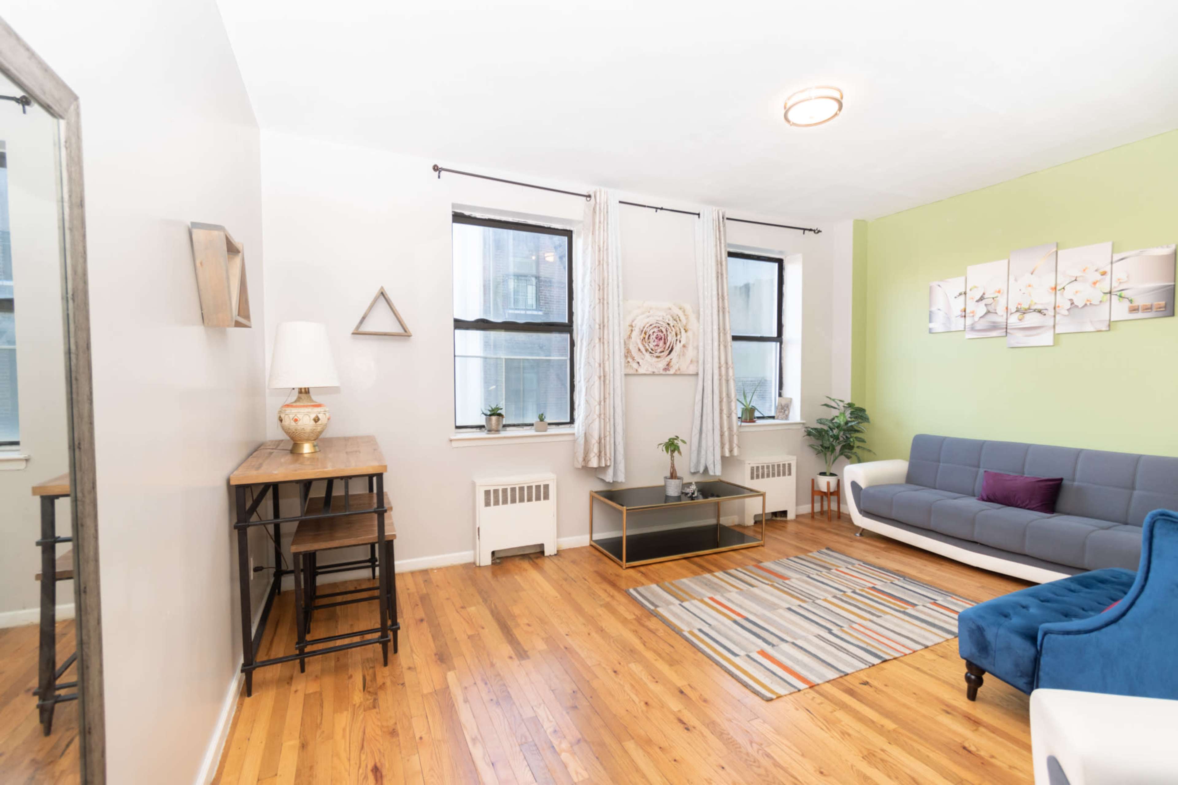 The image shows a living room featuring a blue sofa, a wooden table, large windows, and hardwood flooring.
