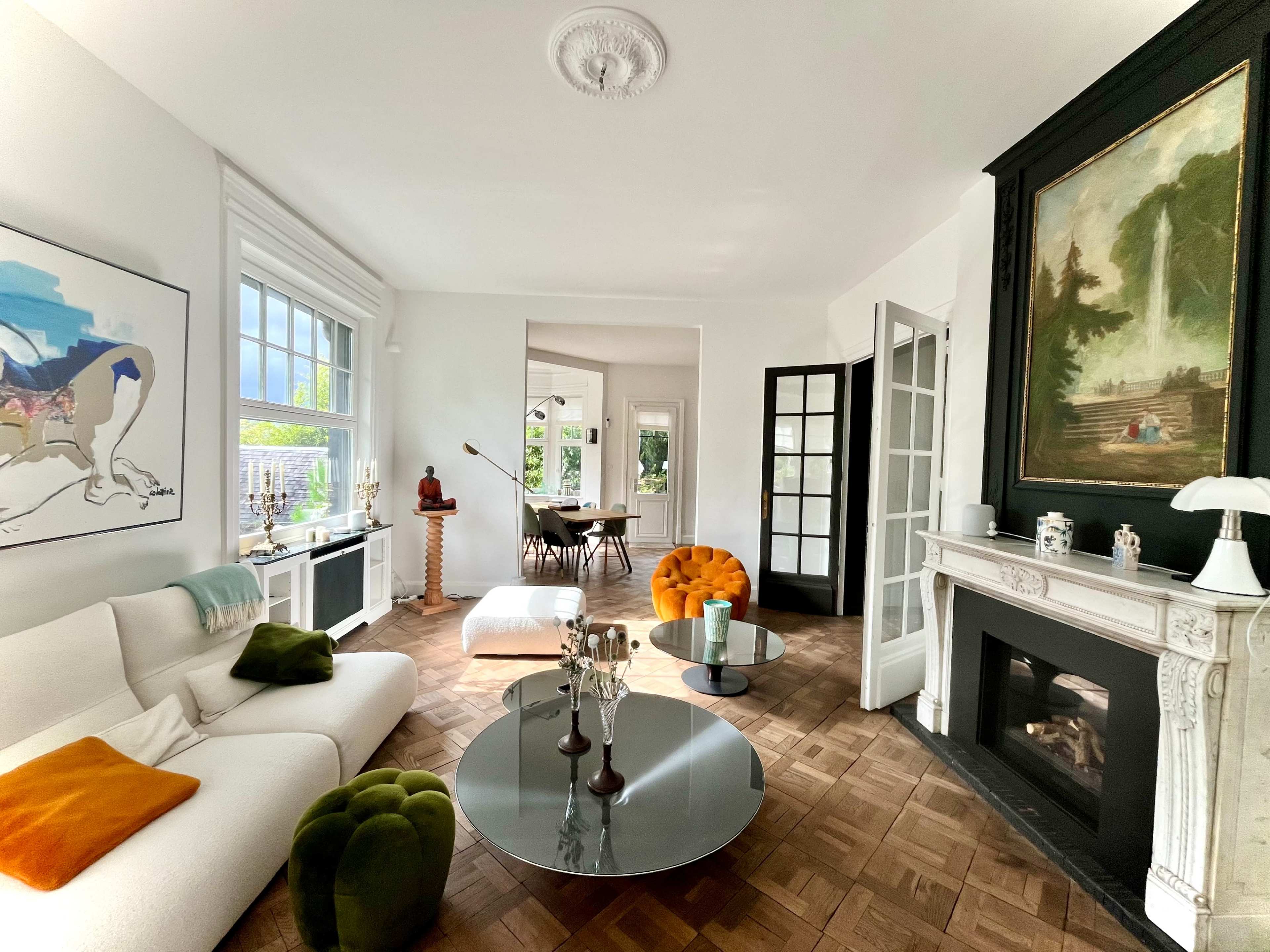 A bright living room features a white sofa with colorful cushions, a round coffee table, and a fireplace, with a dining area visible in the background.