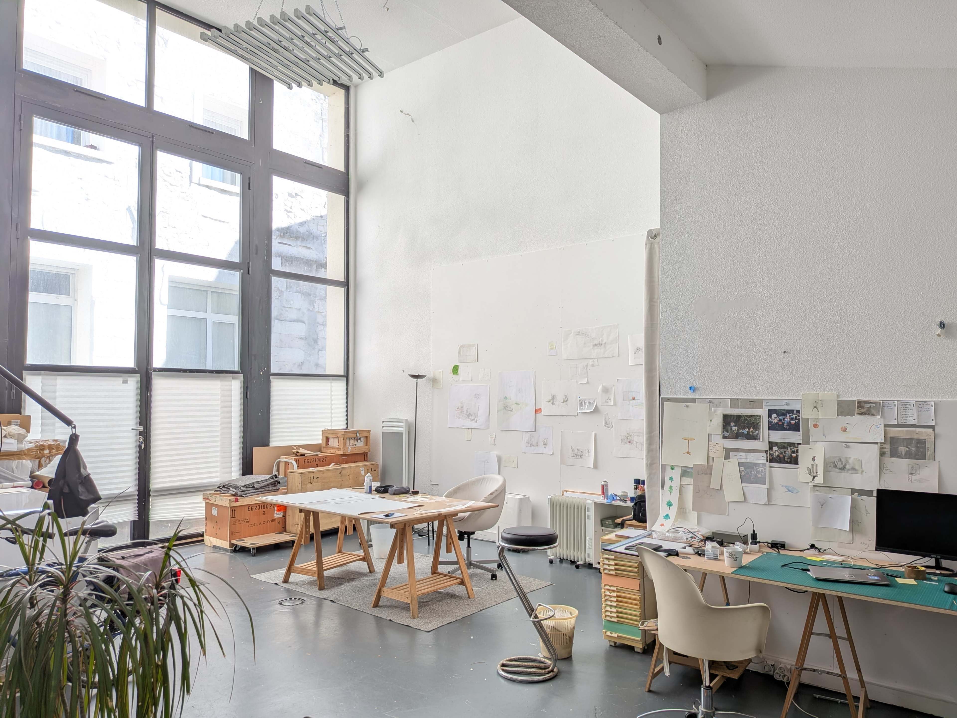 The image depicts a bright studio space with large windows, featuring a workspace with tables, chairs, and walls covered in sketches and documents.