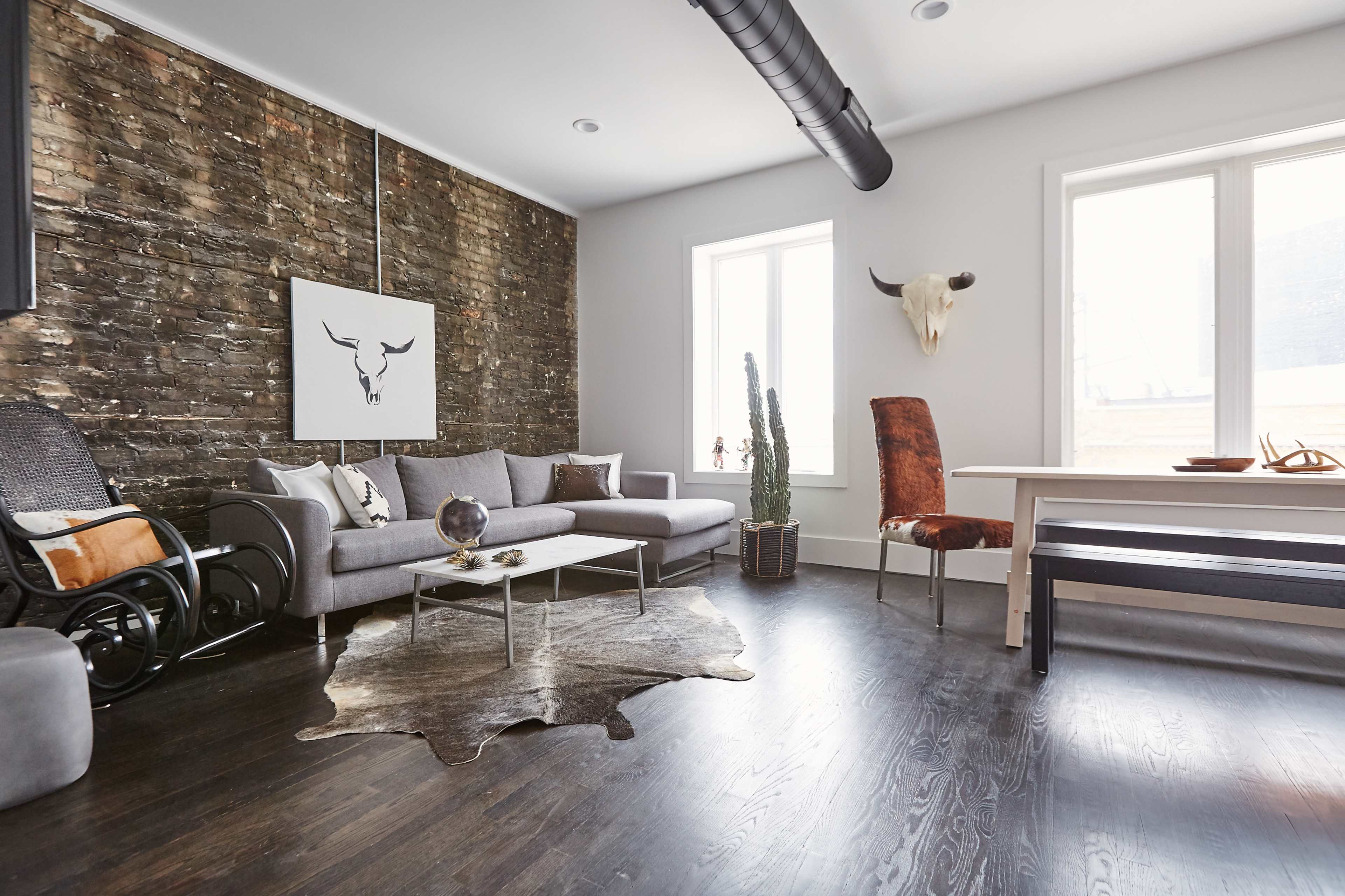 The room features a gray sofa, a wooden table, and a mix of modern and rustic decor against a backdrop of exposed brick walls.