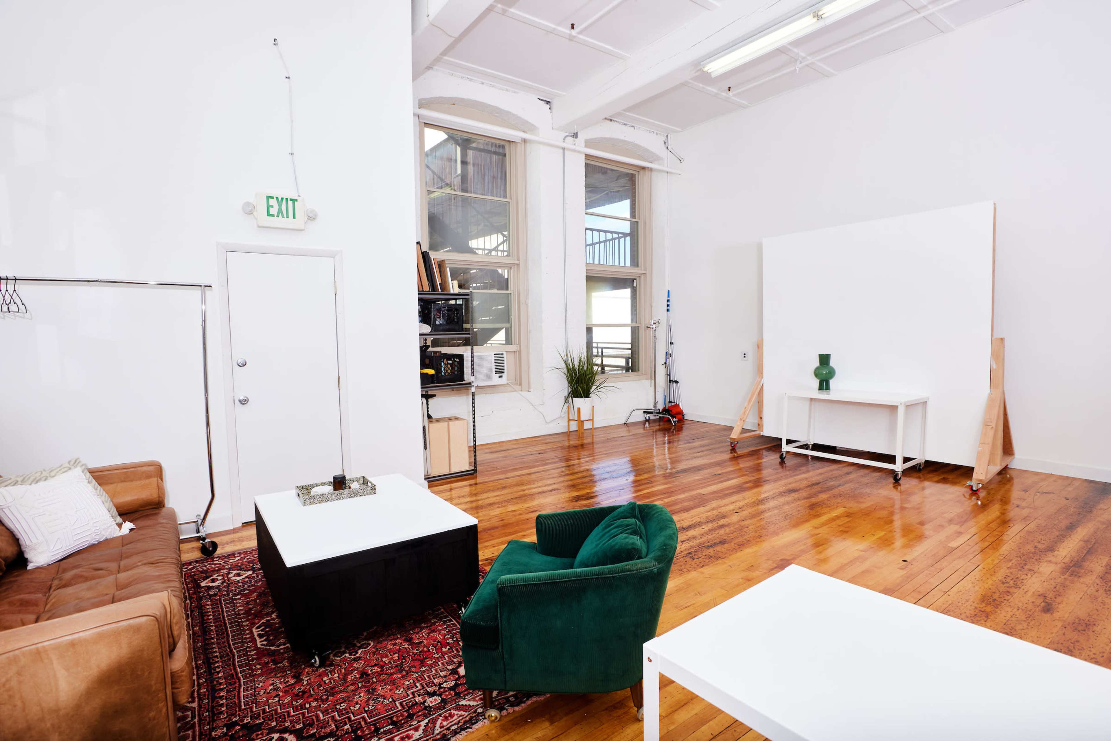 The image shows a spacious, brightly lit room with hardwood floors, featuring a brown leather sofa, a green chair, a white table, and a large blank wall, suggesting a versatile space.