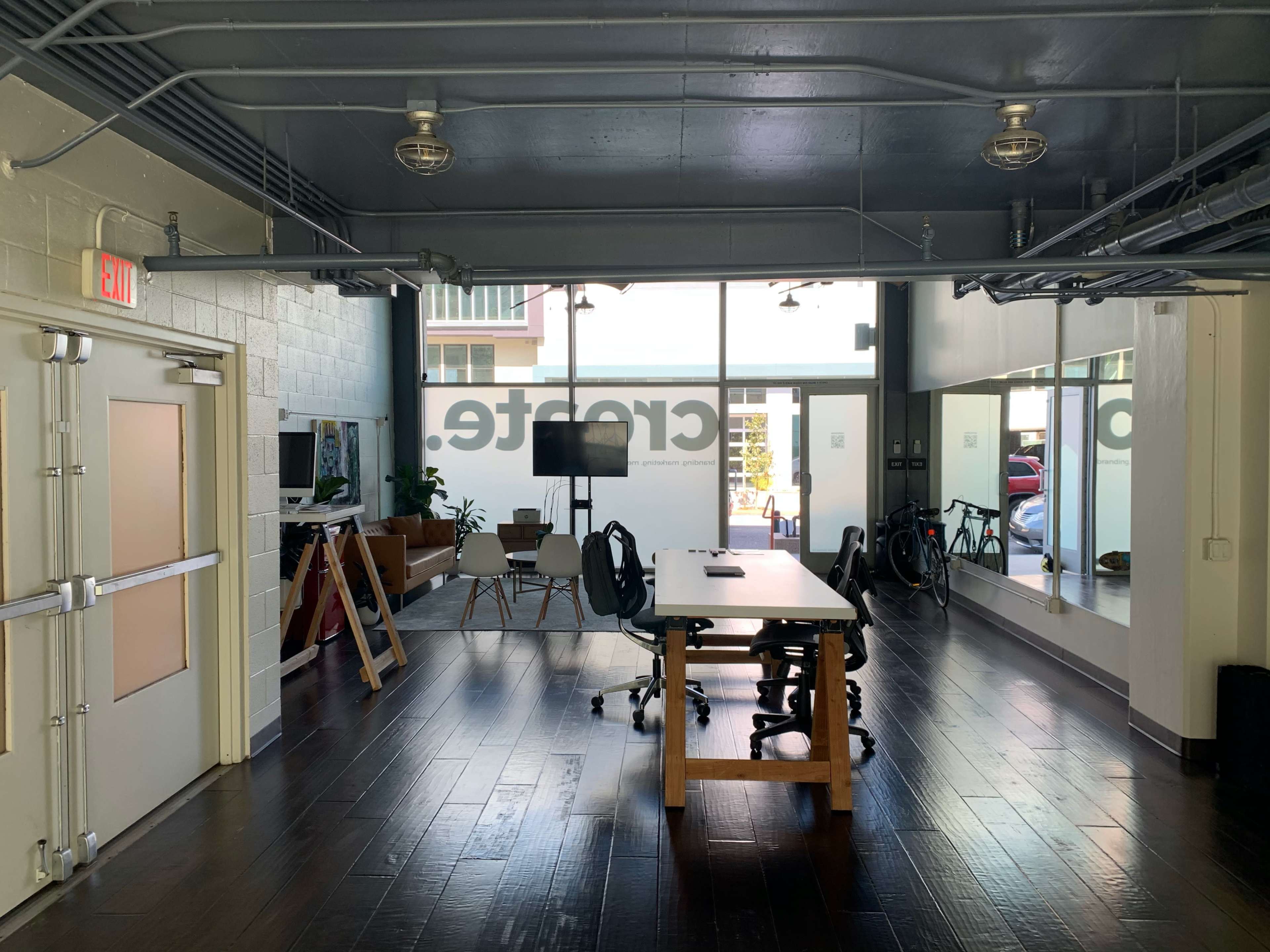 A modern office space with a large table, chairs, and a television, illuminated by natural light from large windows.