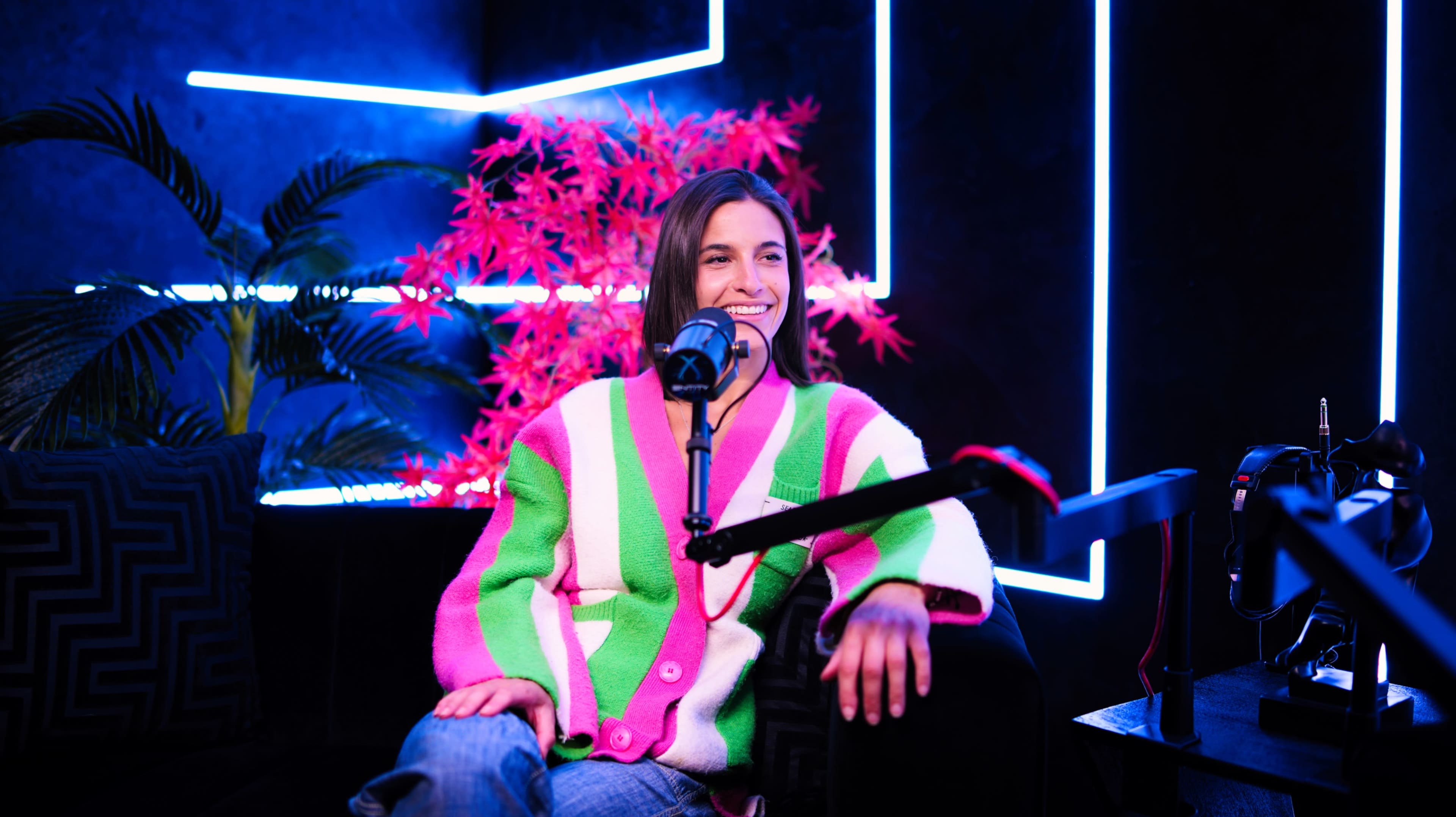 A woman sits on a couch in a podcast studio, wearing a colorful oversized sweater, with neon lights and decorative plants in the background.