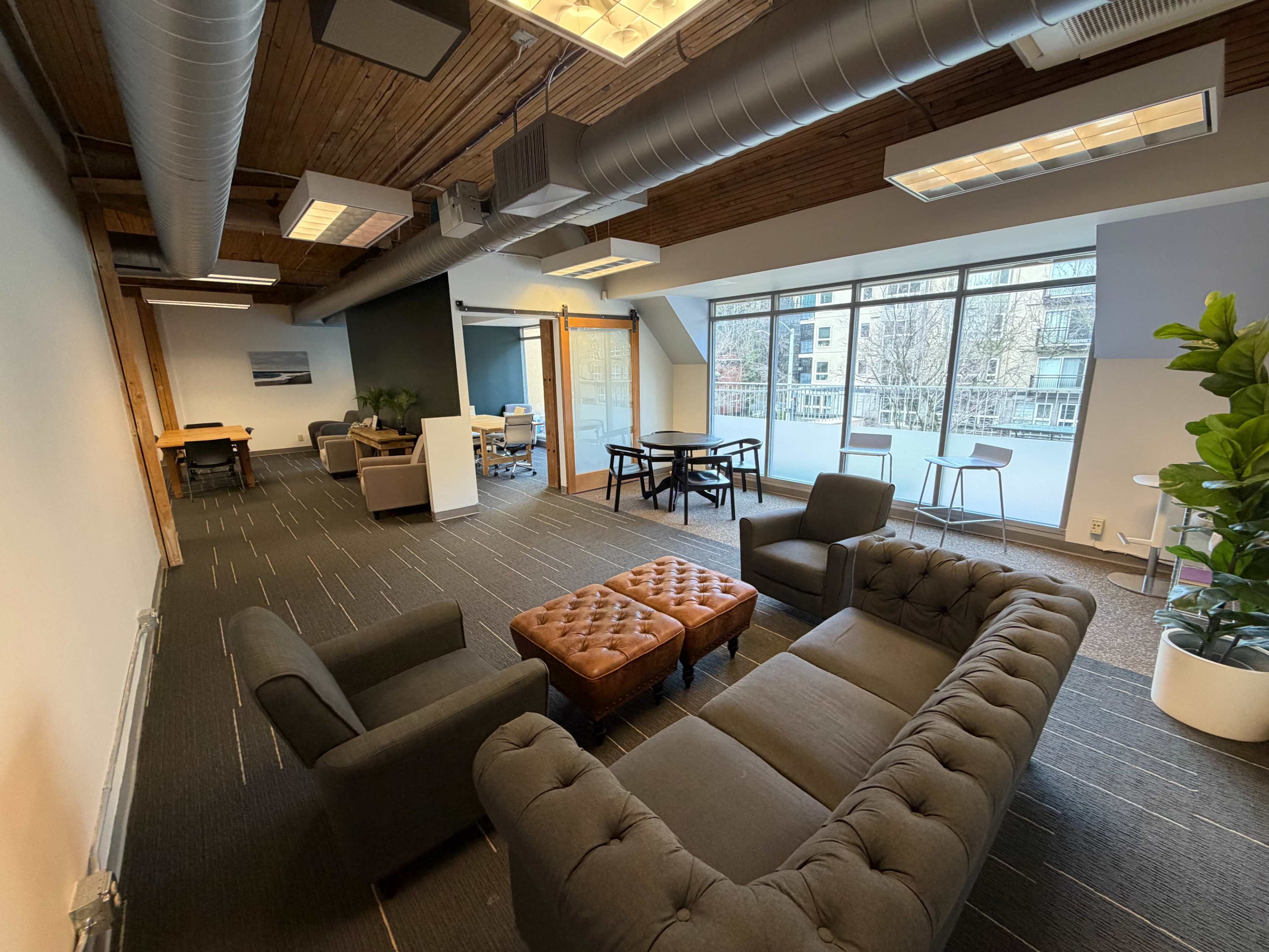The image shows a modern office space with a dark sectional sofa, a tufted ottoman, a table, and large windows allowing natural light to enter.