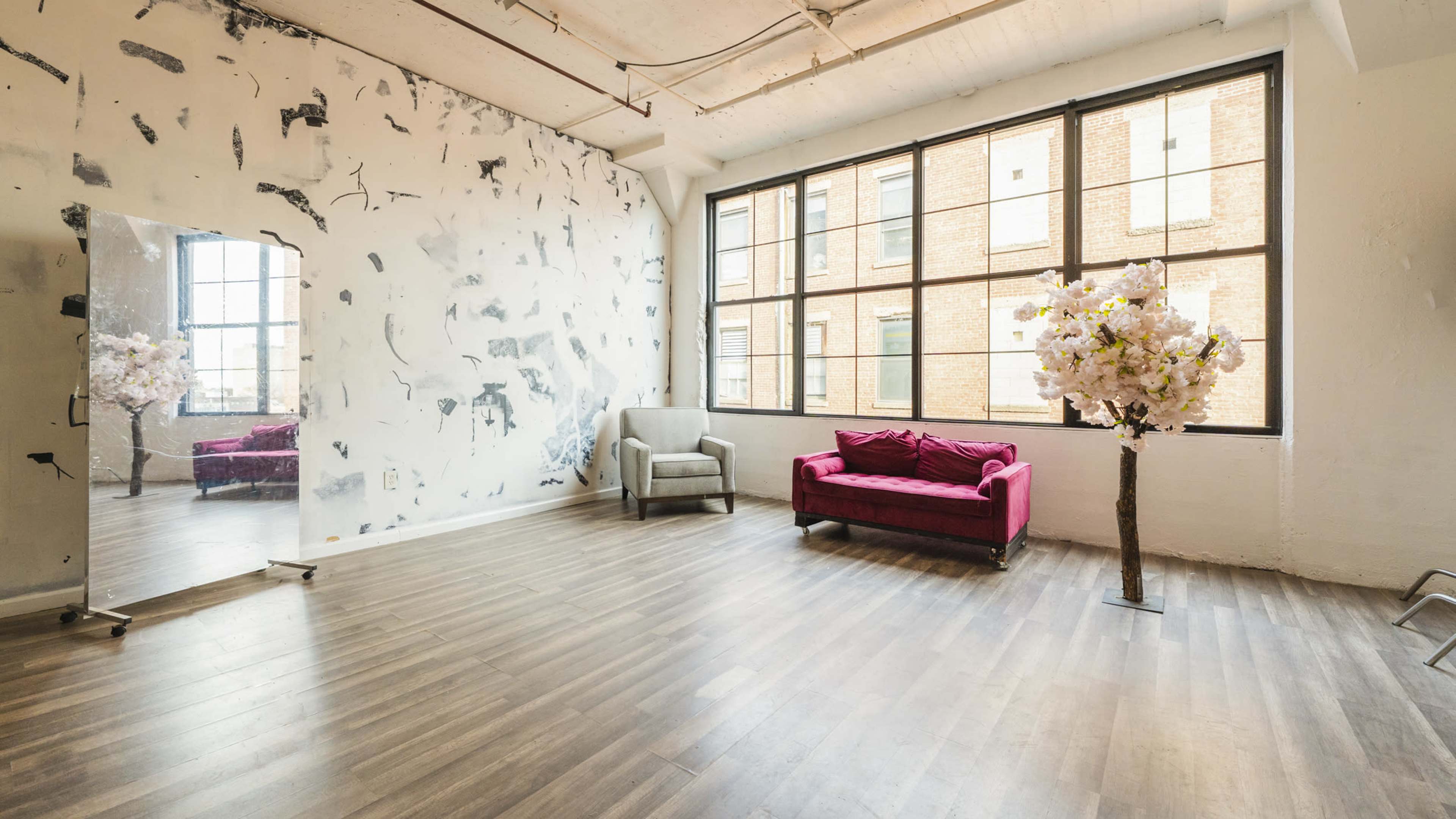A spacious room with large windows, a pink sofa, a gray chair, a mirror, and a decorative tree with pink flowers against a textured white wall.