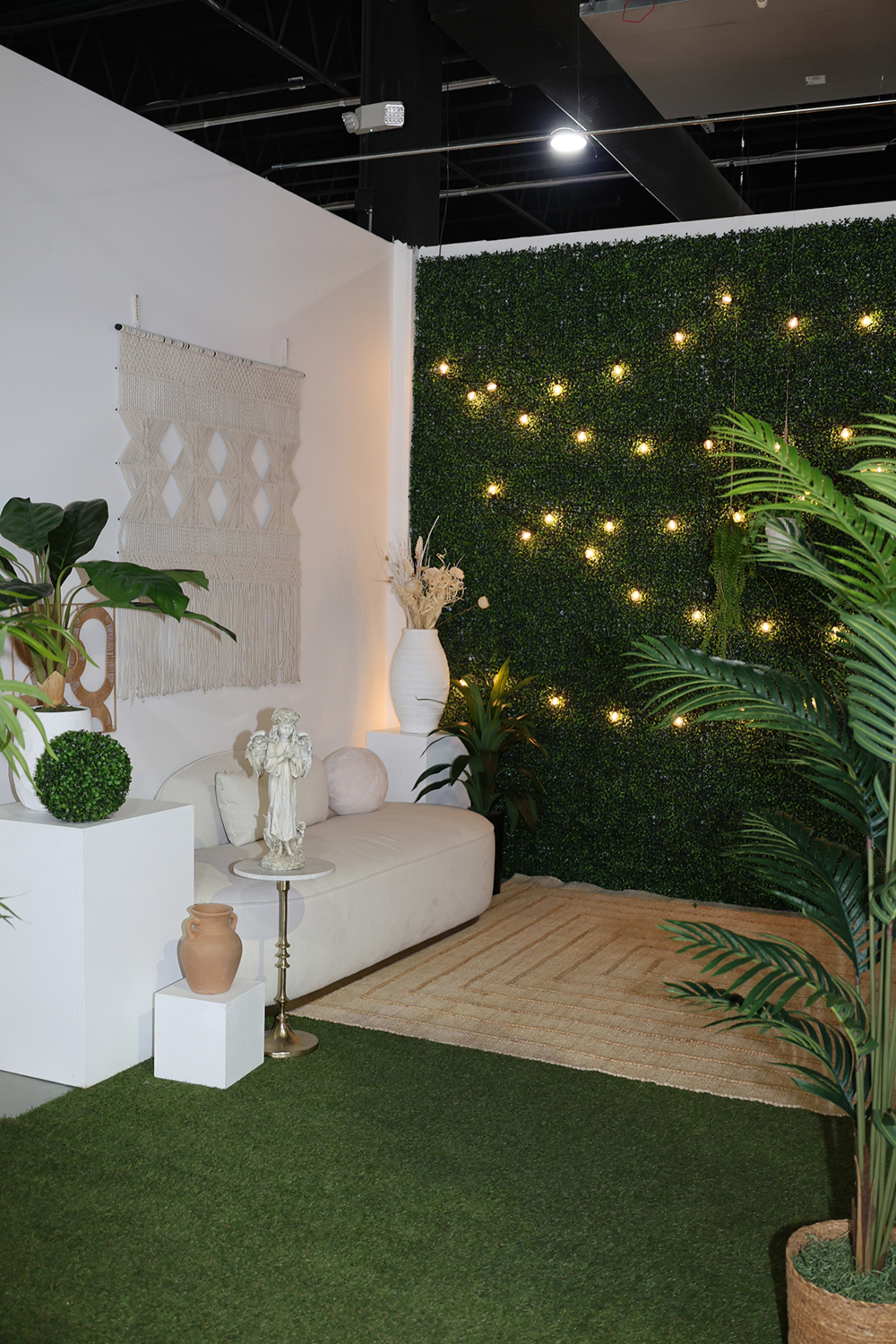 The image features a decorated indoor space with a white sofa, decorative plants, a small table, and a backdrop of a green wall adorned with fairy lights.