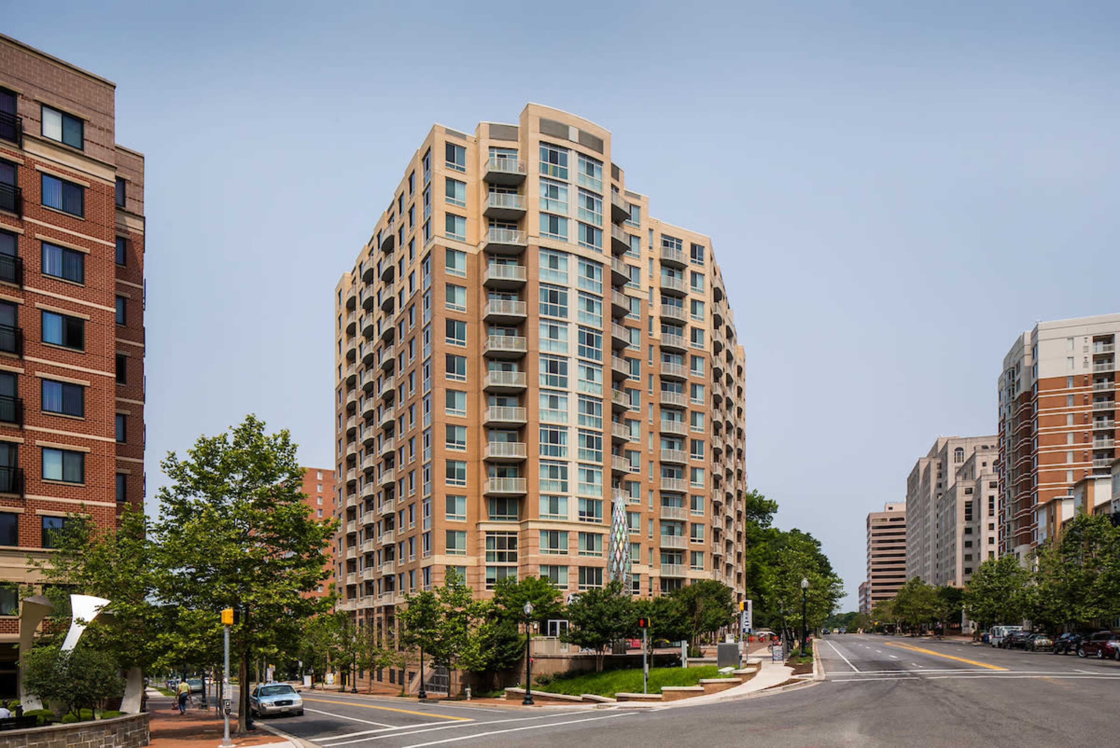 A modern high-rise apartment building stands at the corner of a city intersection, surrounded by tree-lined streets and other buildings.