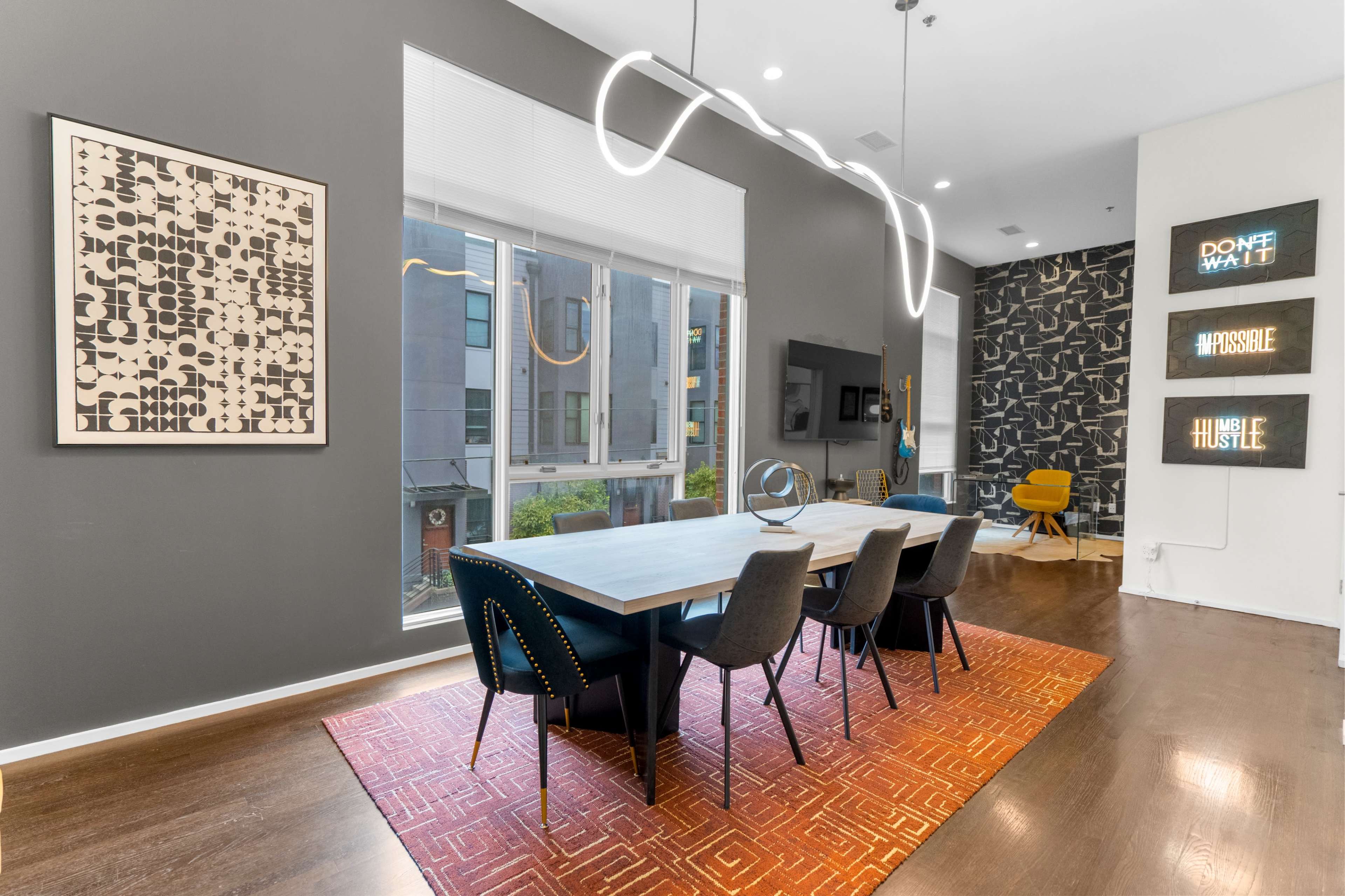A modern dining area features a long table surrounded by upholstered chairs, set against a backdrop of large windows and creatively designed wall decor.
