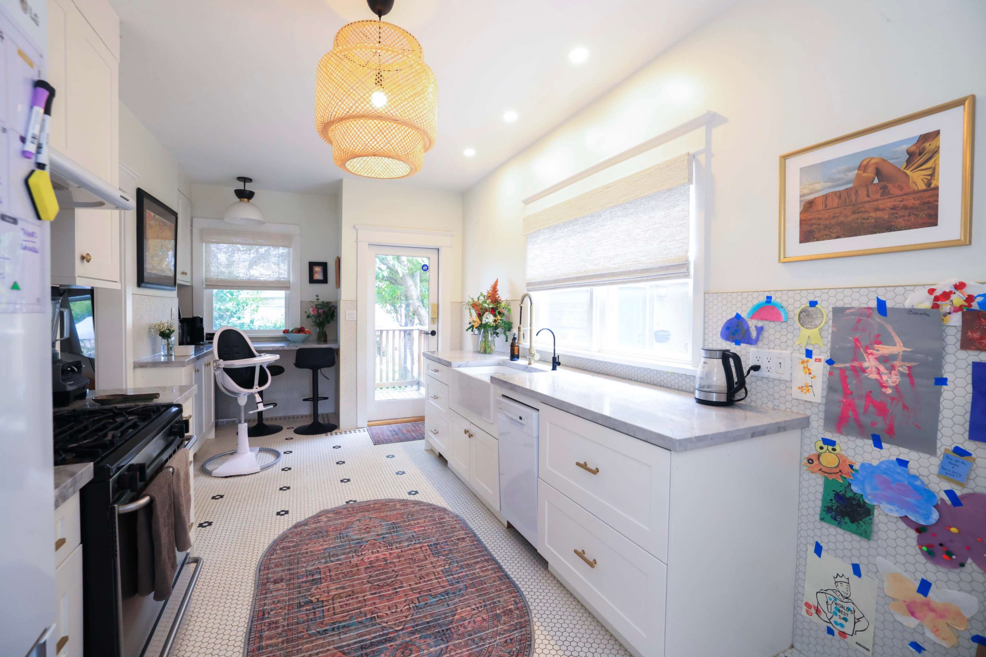 The image shows a bright kitchen featuring white cabinetry, a large window with shades, and a dining area with a bar stool.