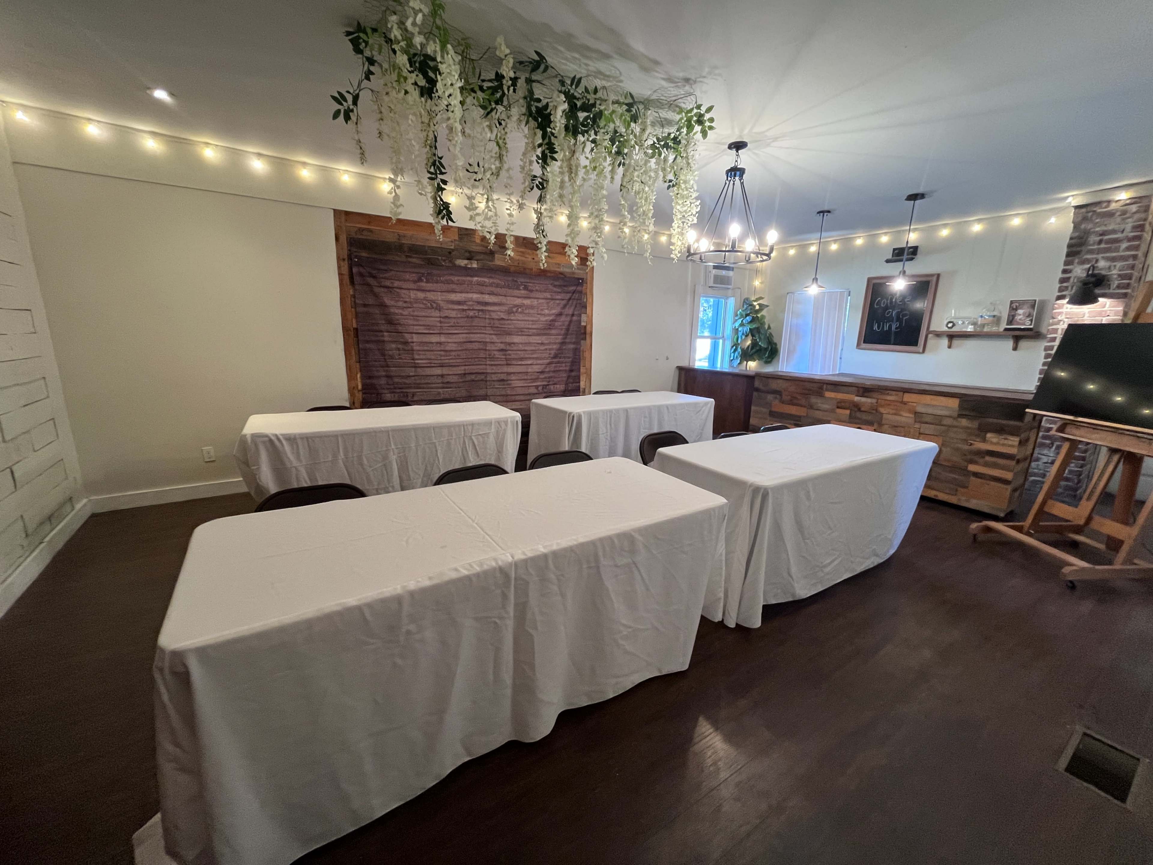 The image shows a small, well-lit event space with five covered tables arranged in rows, surrounded by decorative elements such as plants and string lights.