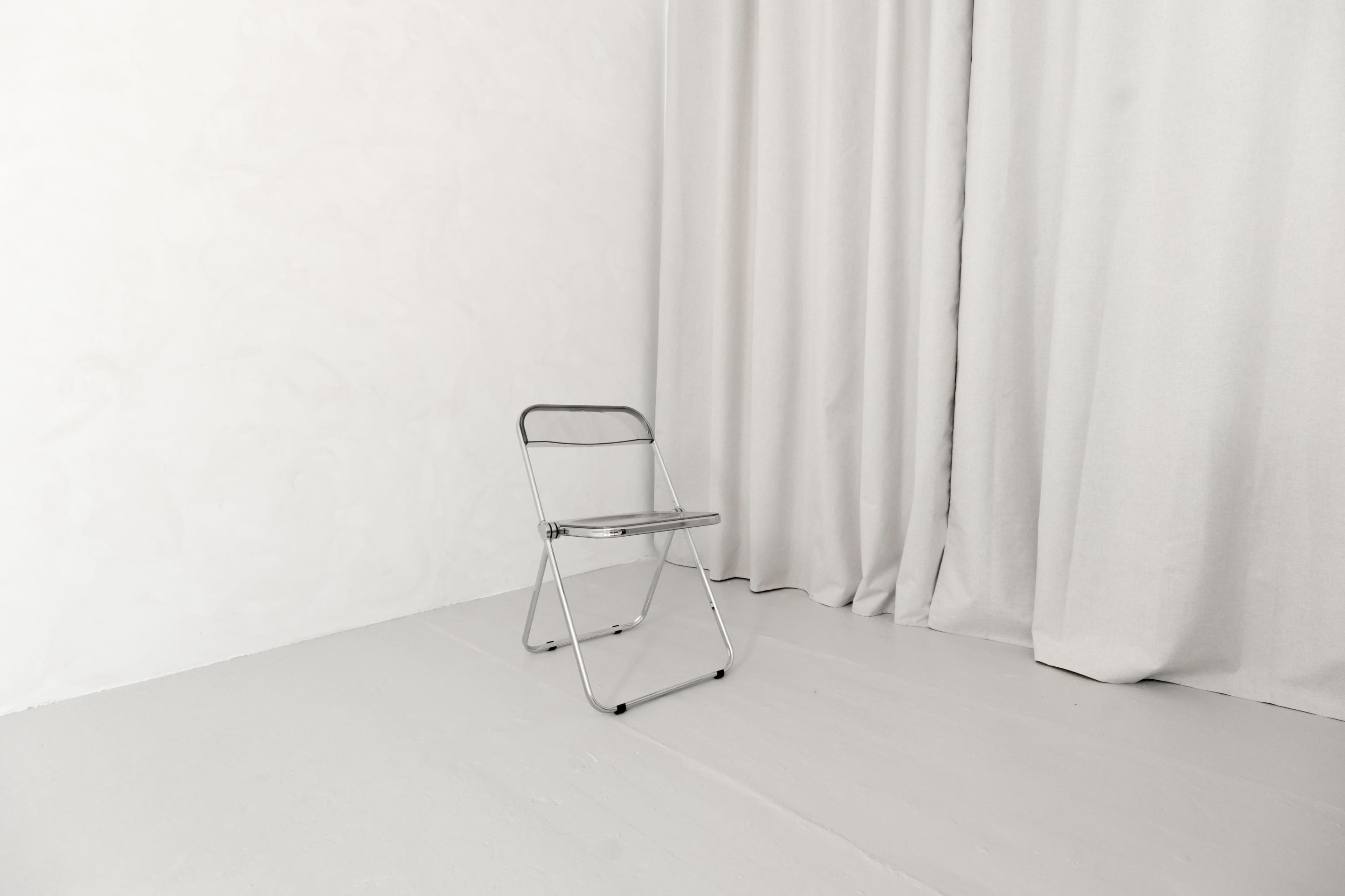 A folding metal chair is positioned in a sparse, minimalist room with a light-colored floor and draped fabric curtains.