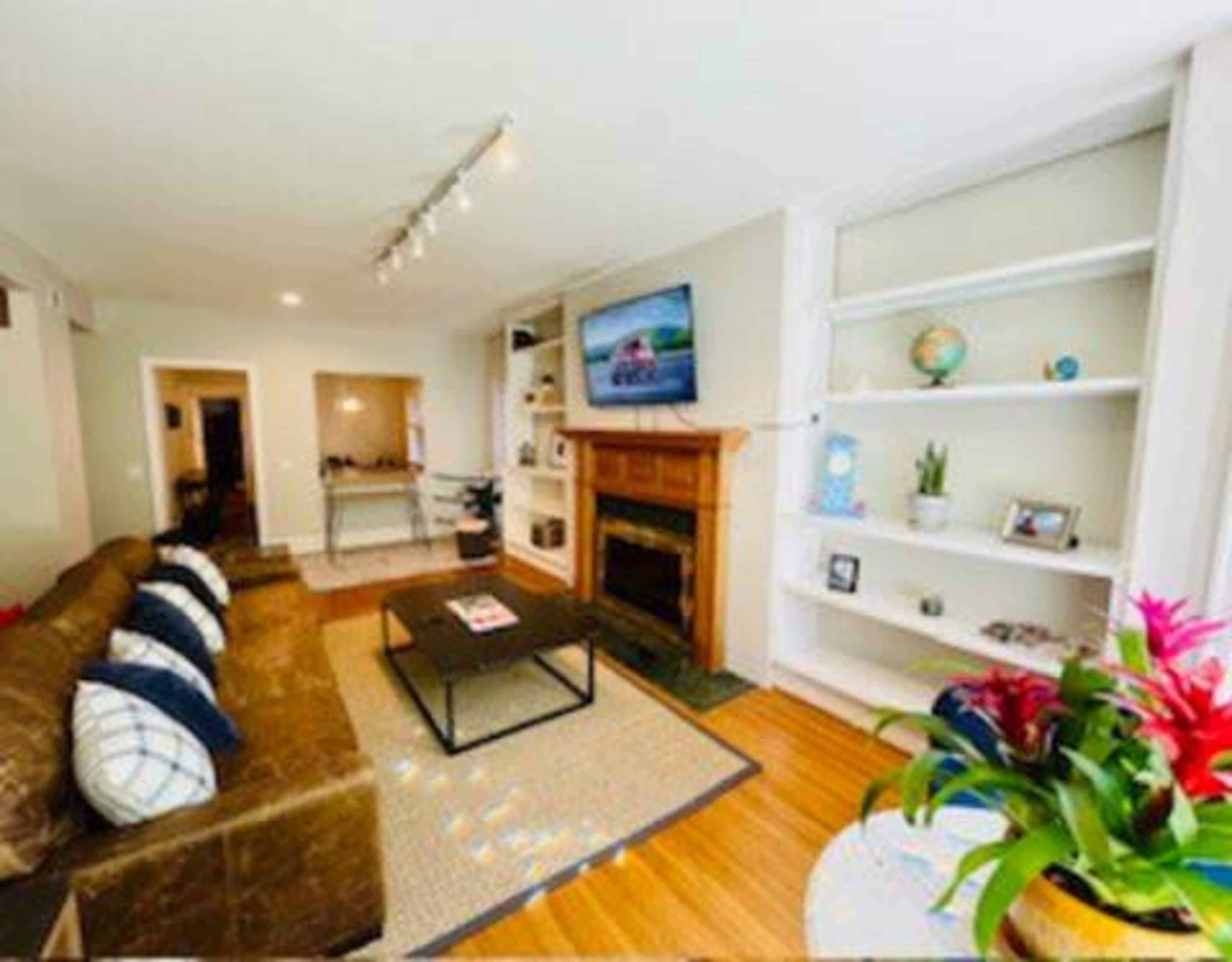 The image shows a living room featuring a brown sofa, a coffee table, a TV mounted above a wooden mantle, and built-in shelves with decorative items and plants.