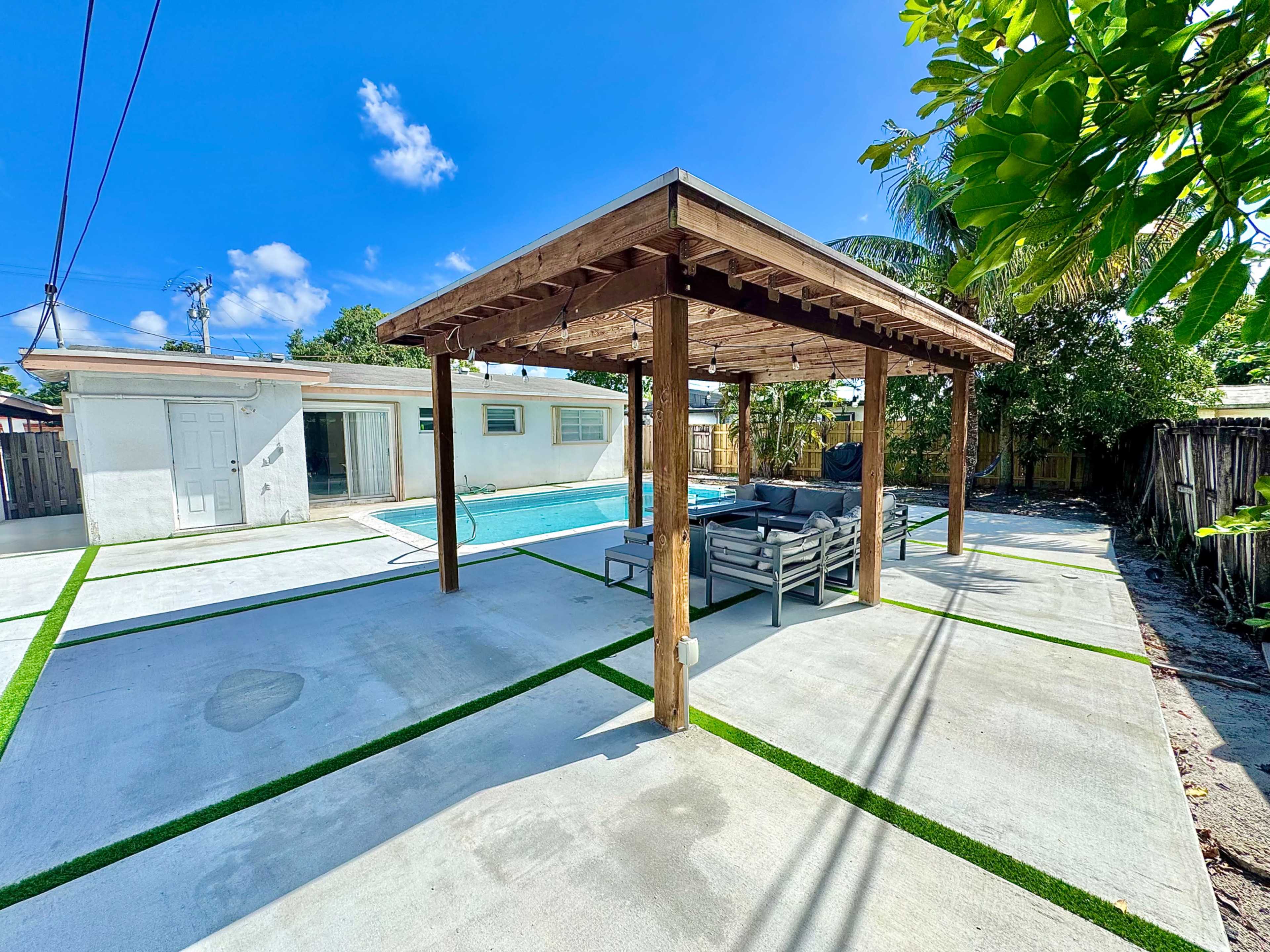 The image shows a patio area with a covered structure, outdoor seating, and a swimming pool in the background, surrounded by well-maintained landscaping.