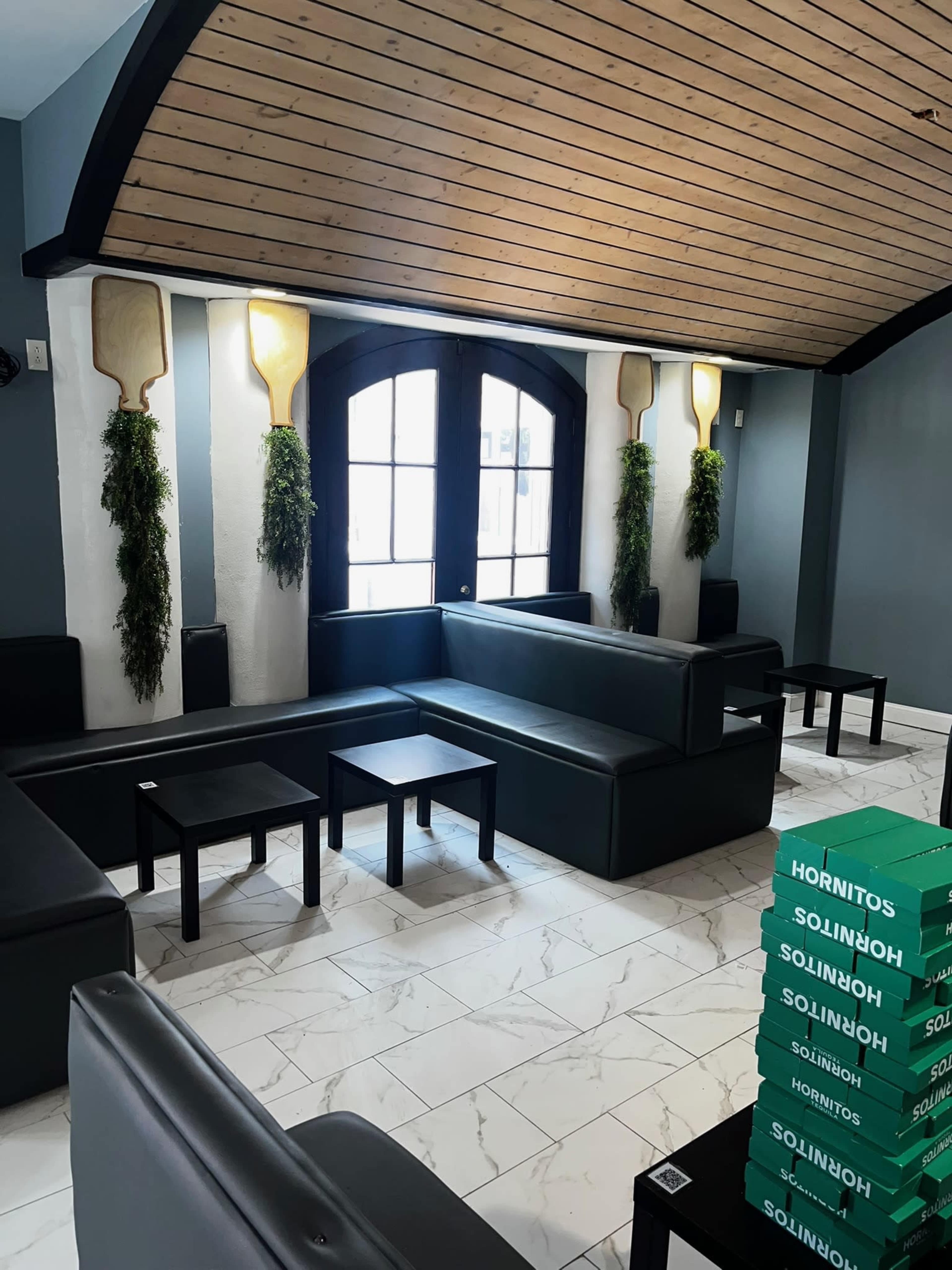The image shows a modern lounge area with black sofas and tables, featuring a large window and decorative plants on the walls.