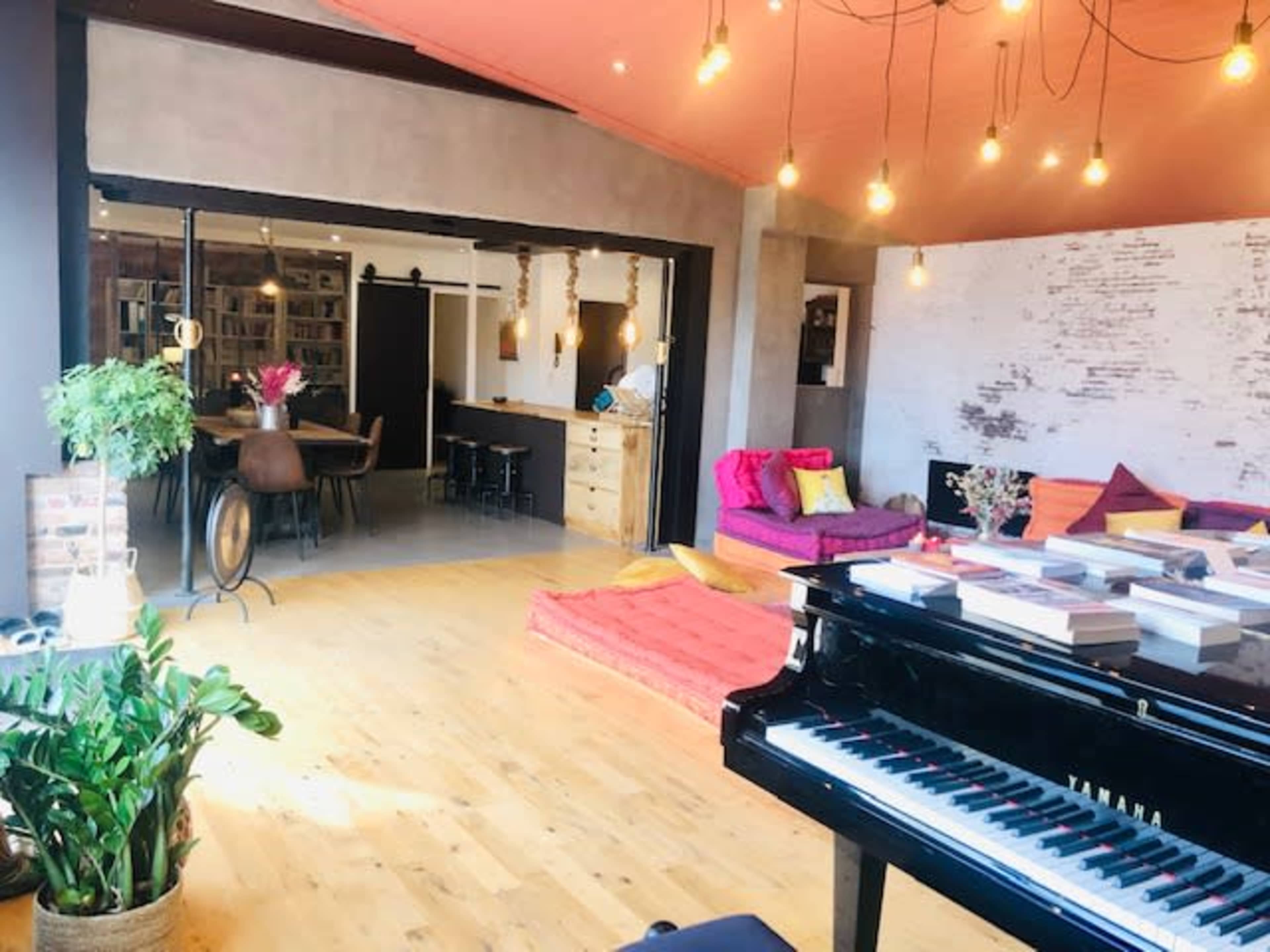 A spacious living area features a grand piano in the foreground, colorful seating arrangements, and an open kitchen in the background.