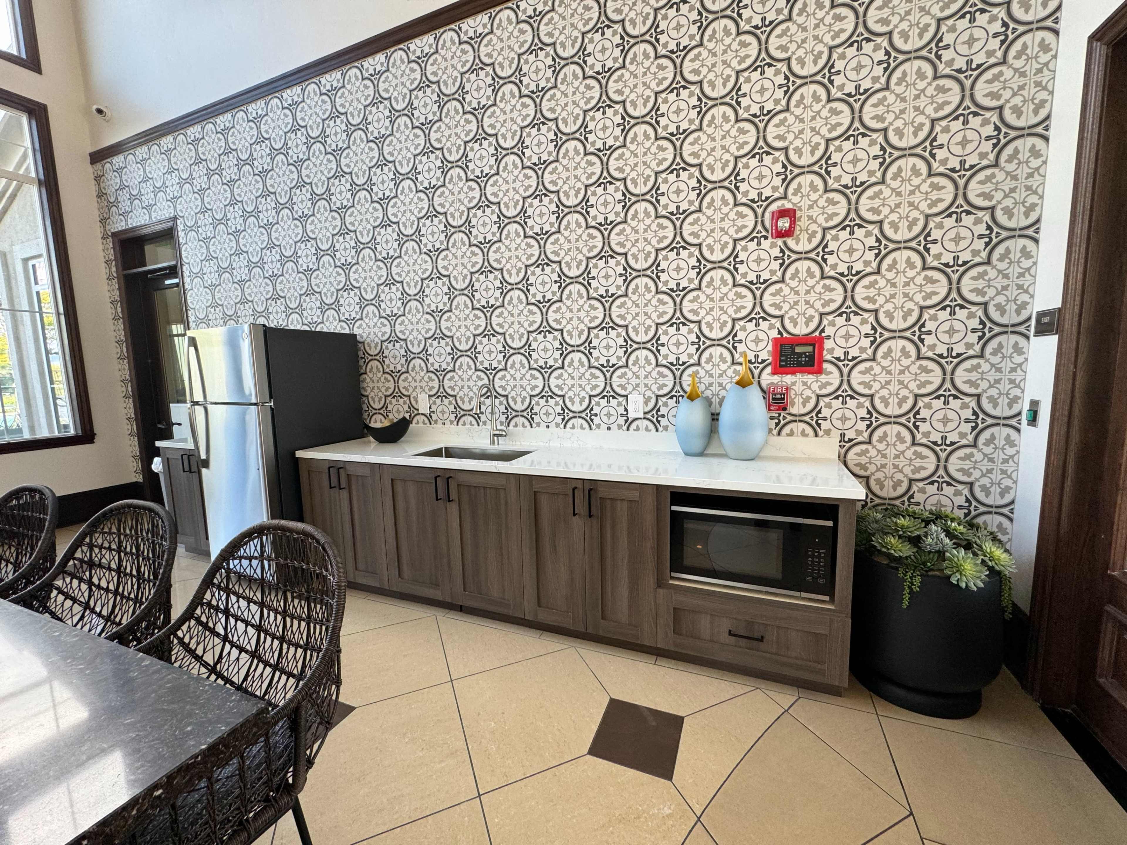 A modern kitchen area featuring a patterned tiled wall, a stainless steel refrigerator, a countertop with a sink, and decorative vases on display.