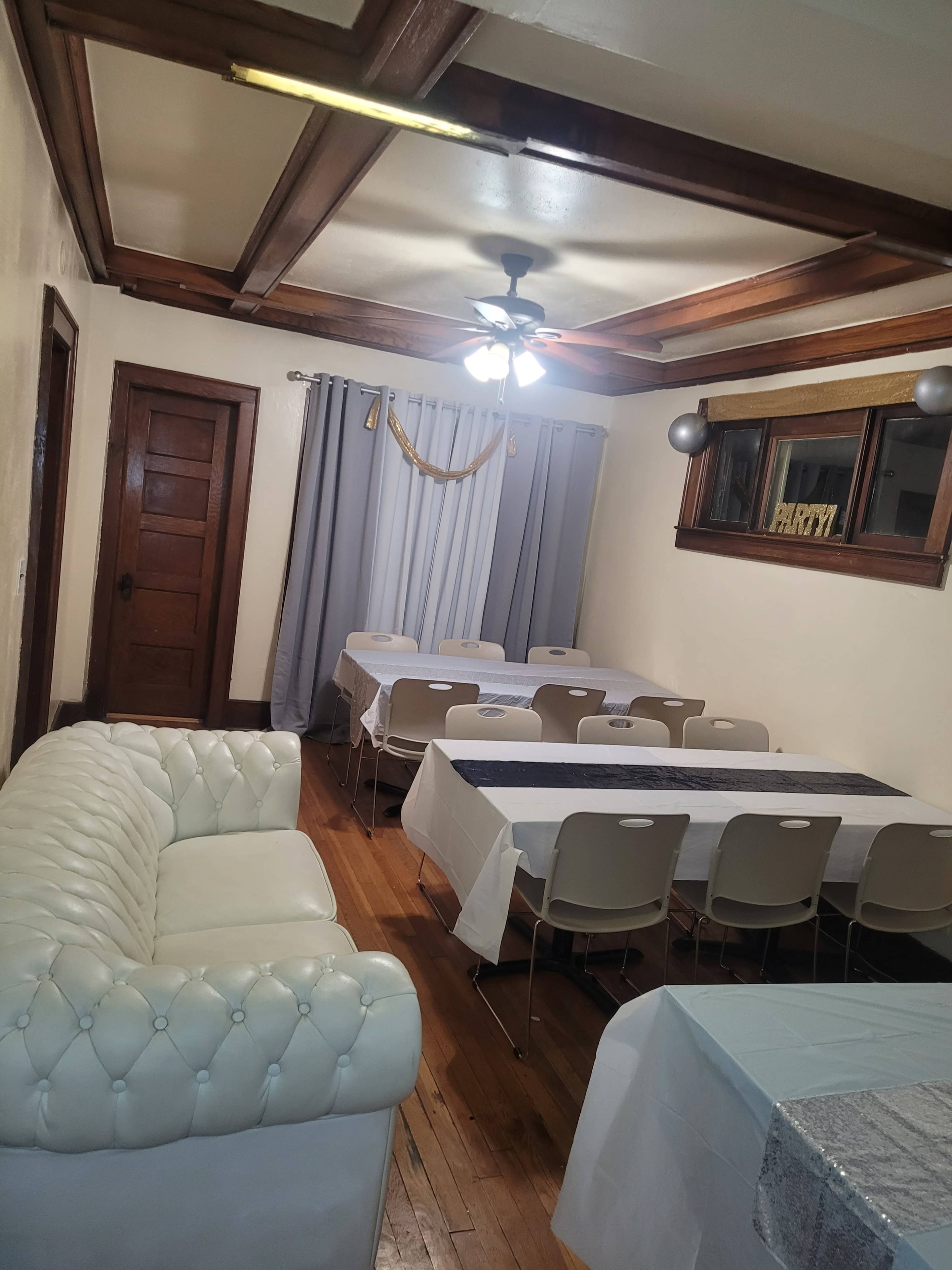 A room with wooden floors, a white leather sofa, several tables covered with white tablecloths, and curtains at the windows.