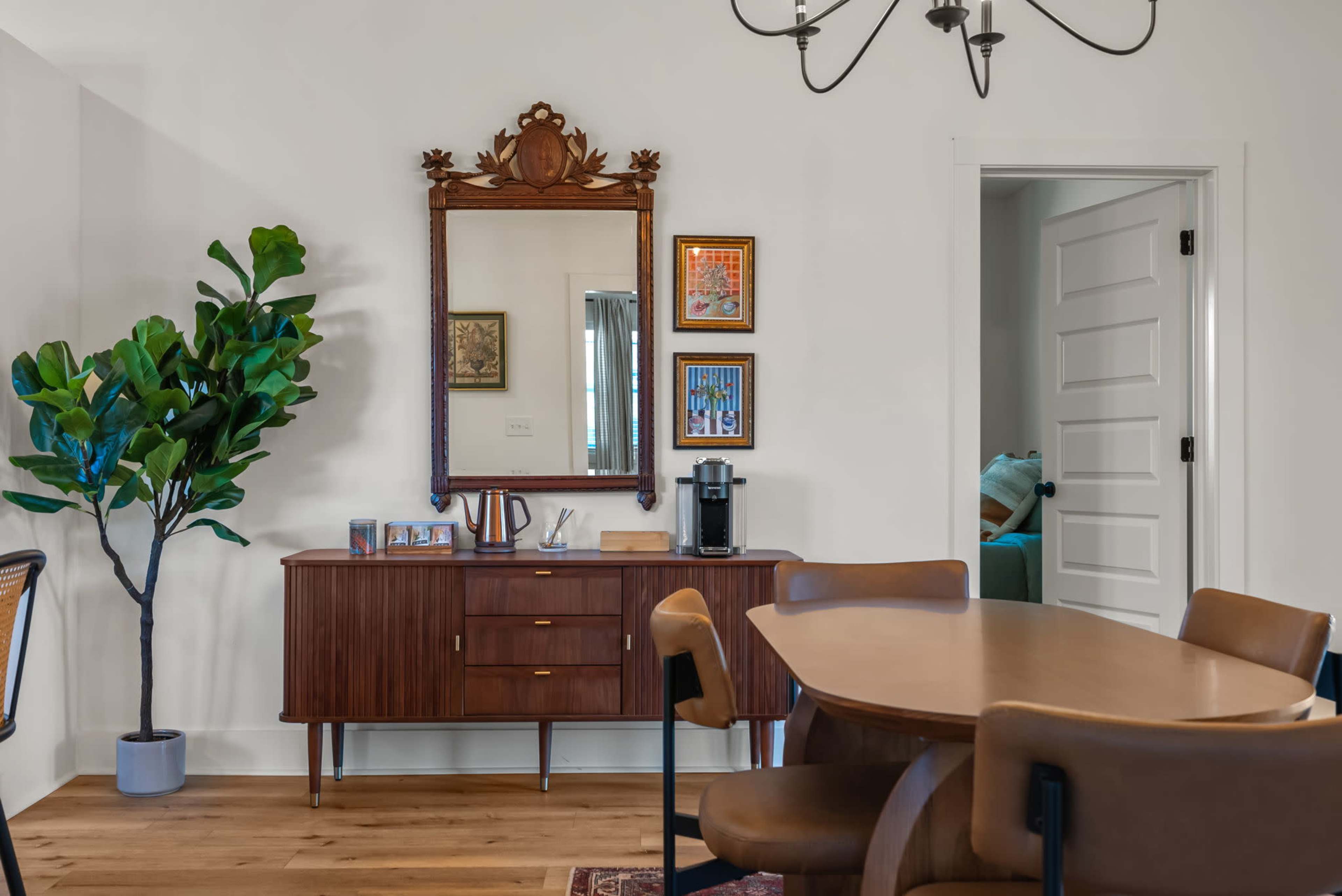 A wood sideboard with a large mirror hangs above it, accompanied by framed artwork and a coffee maker, while a potted plant and a dining table with chairs occupy the space.