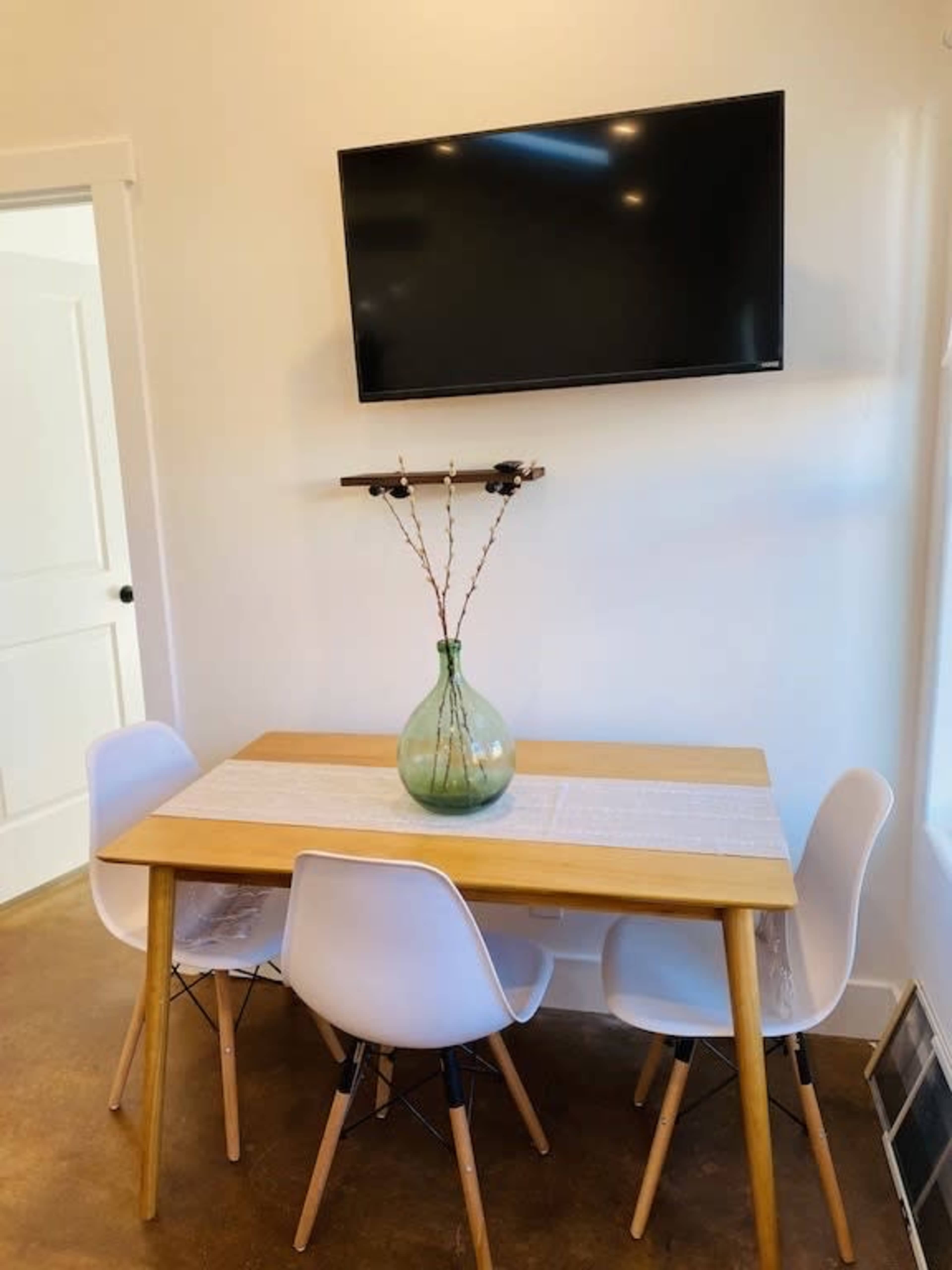 A wooden dining table with four white chairs is set against a wall with a mounted TV above and a vase of branches on the table.