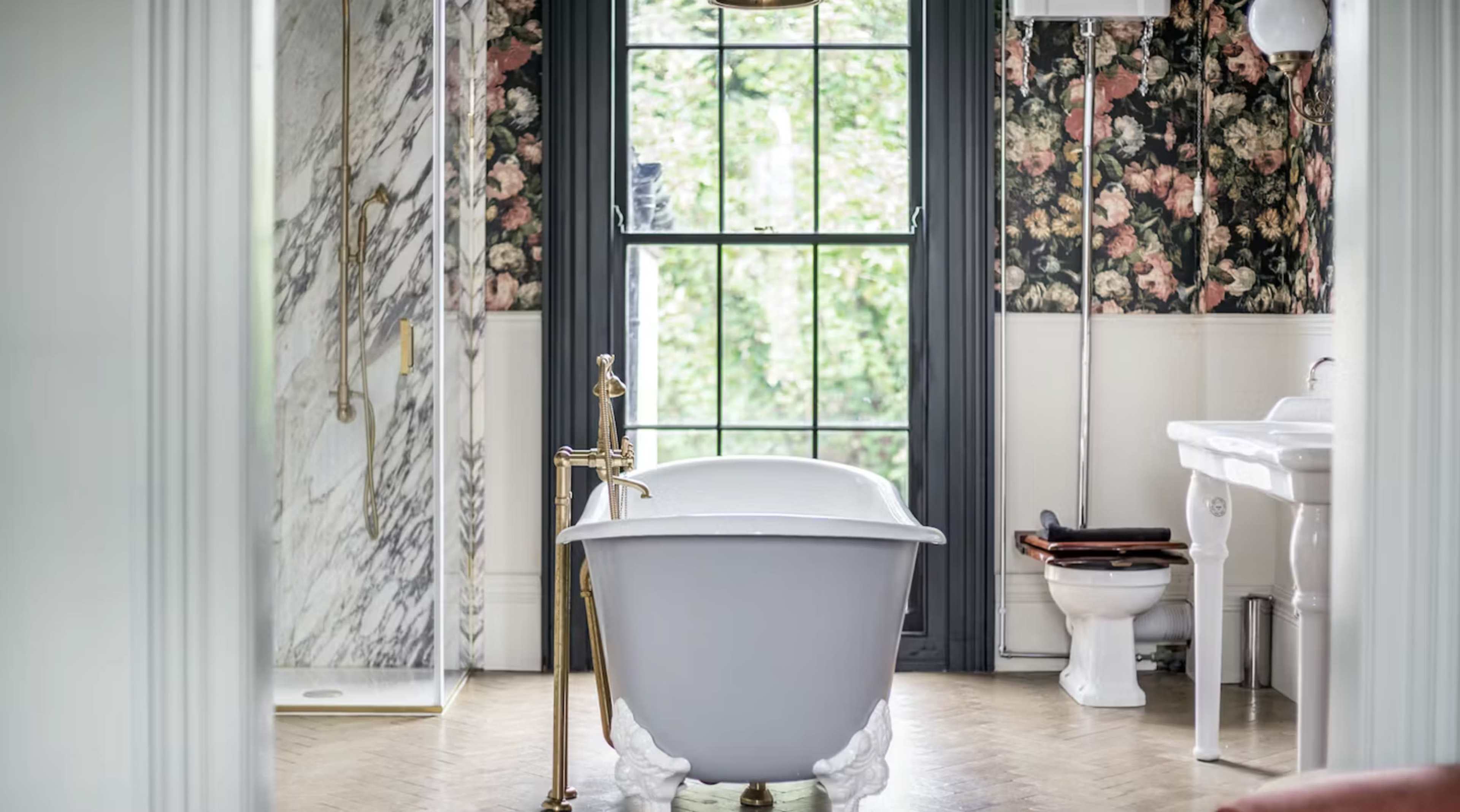A bathroom with a clawfoot bathtub, a shower area with marble walls, and floral wallpaper alongside a window providing natural light.