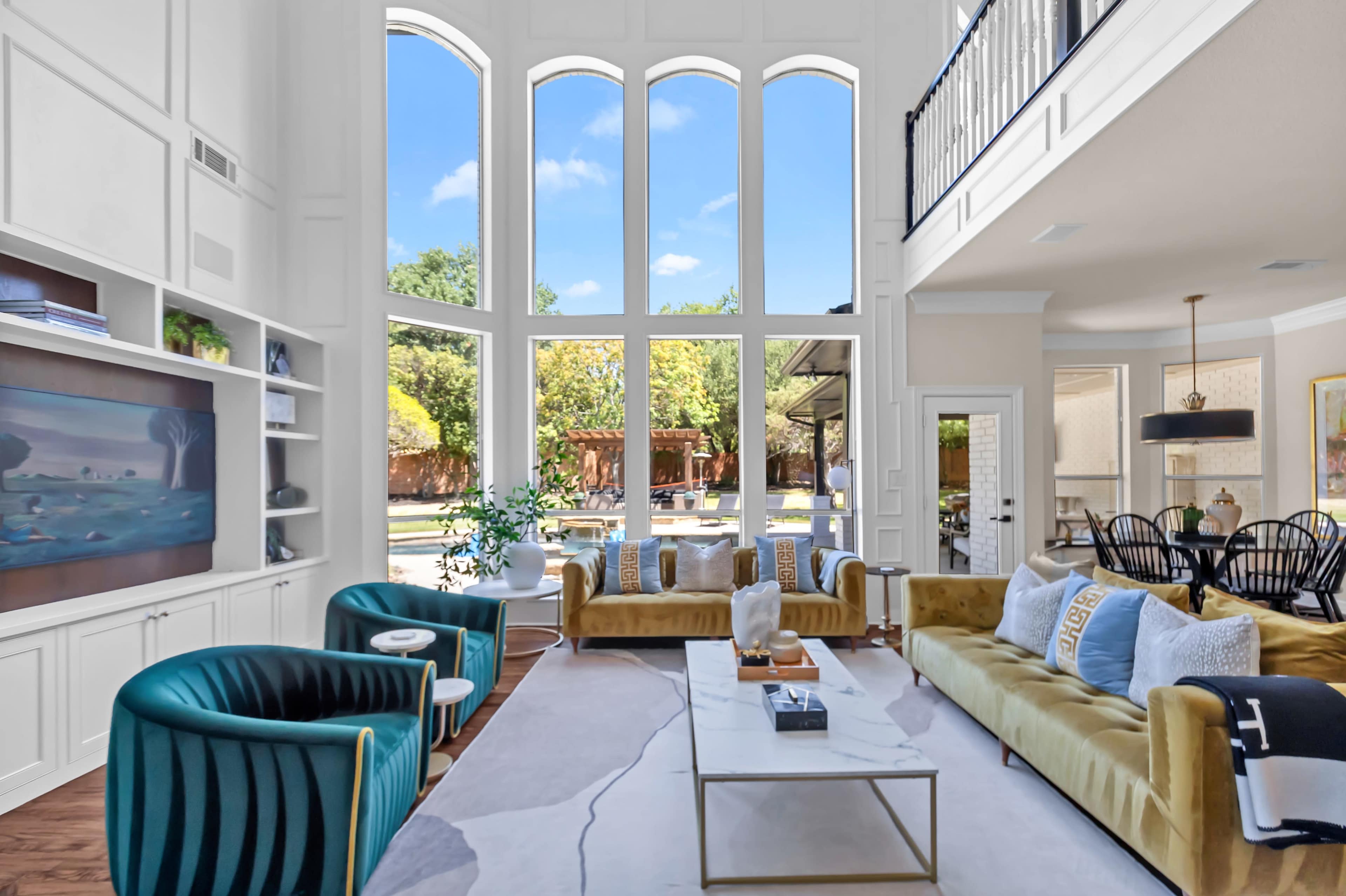 The image shows a bright living room featuring large windows, modern seating arrangements with colorful sofas, and an open view of a backyard patio.