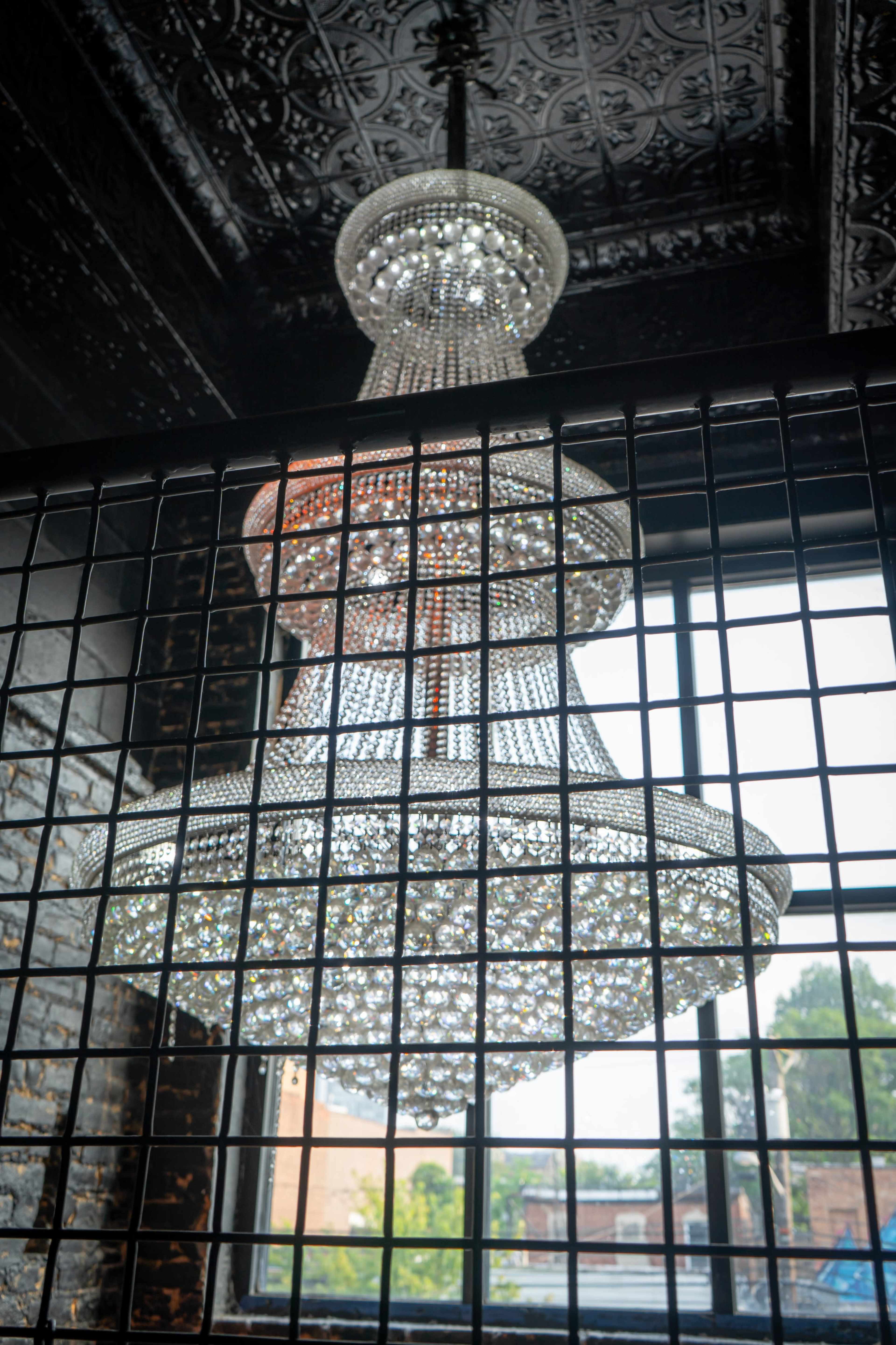 A large crystal chandelier hangs from a high ceiling, partially framed by a metal grid, with a window visible in the background.
