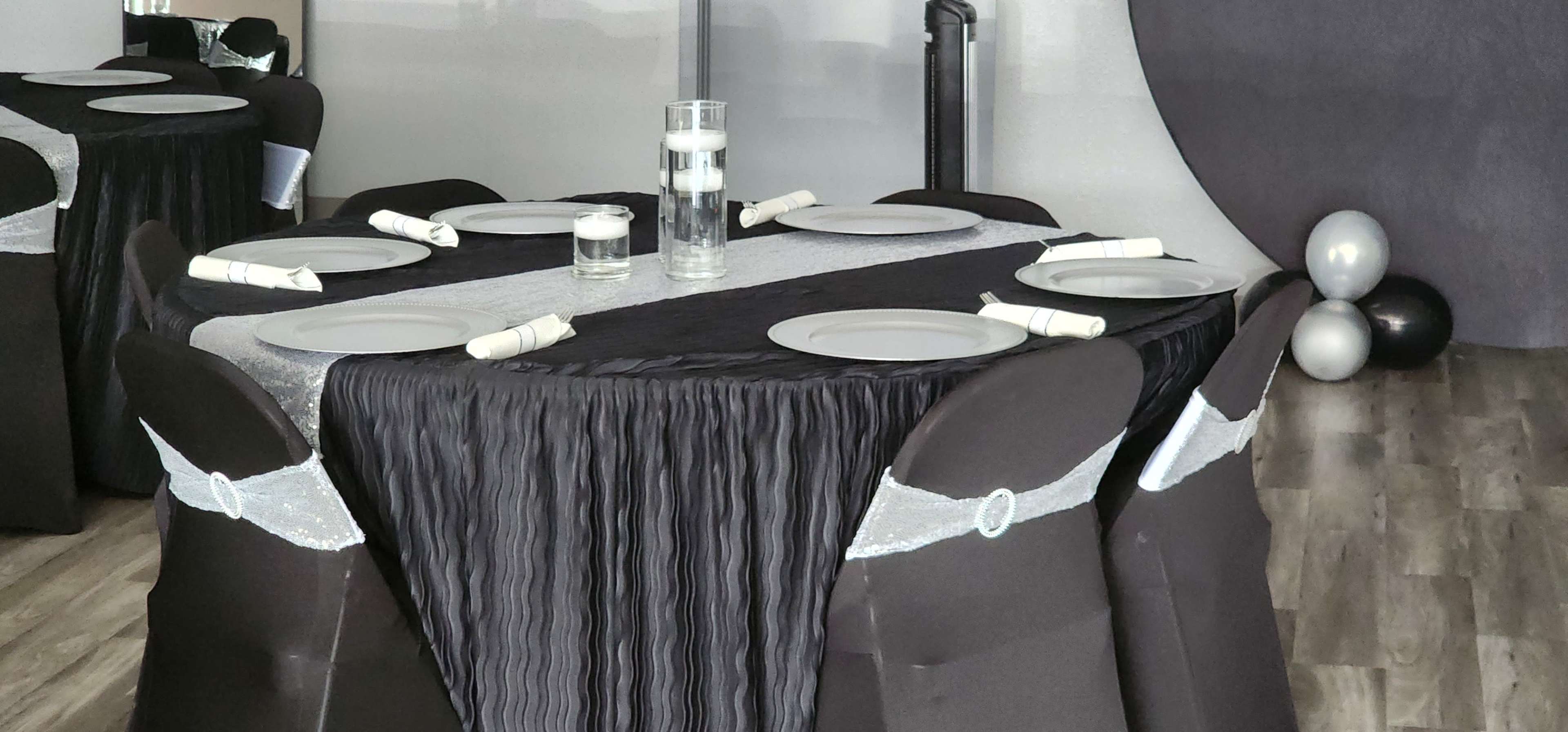 The image shows a neatly set dining table with a black and silver tablecloth, round plates, and stylish black chairs adorned with silver sashes, in a modern event space.