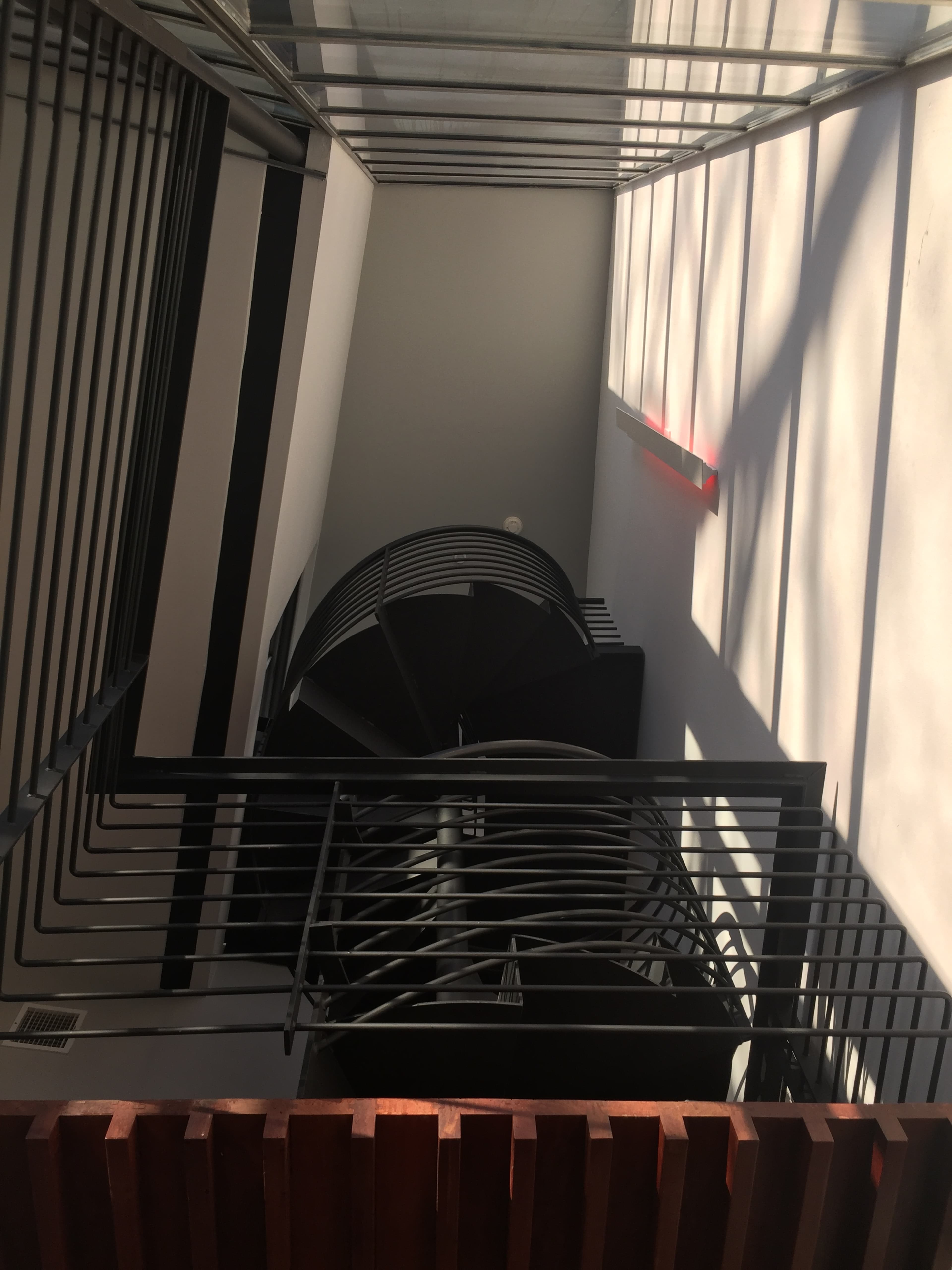 A spiral staircase viewed from above, surrounded by walls and railings casting long shadows.