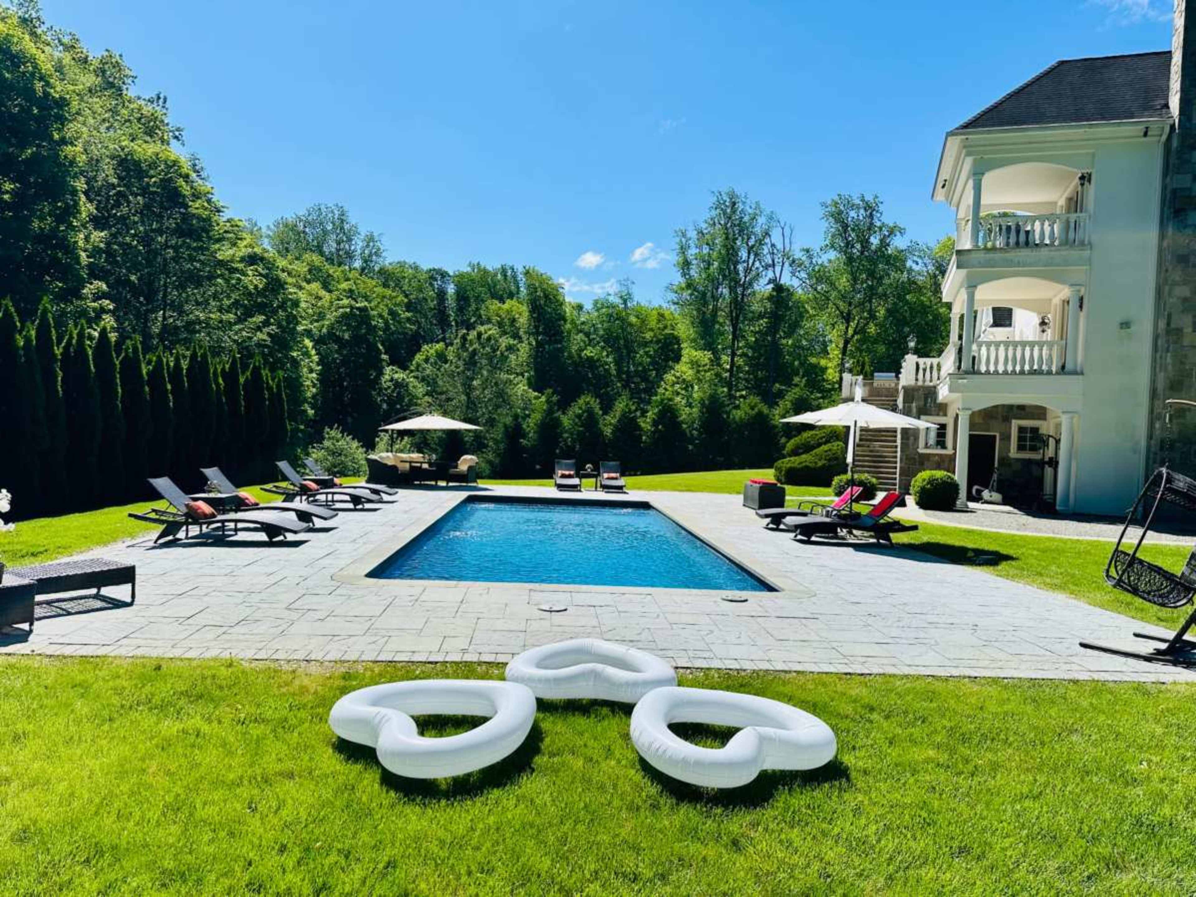 The image shows a spacious backyard with a swimming pool, surrounded by lounge chairs and a manicured lawn, with trees and a multi-story house in the background.