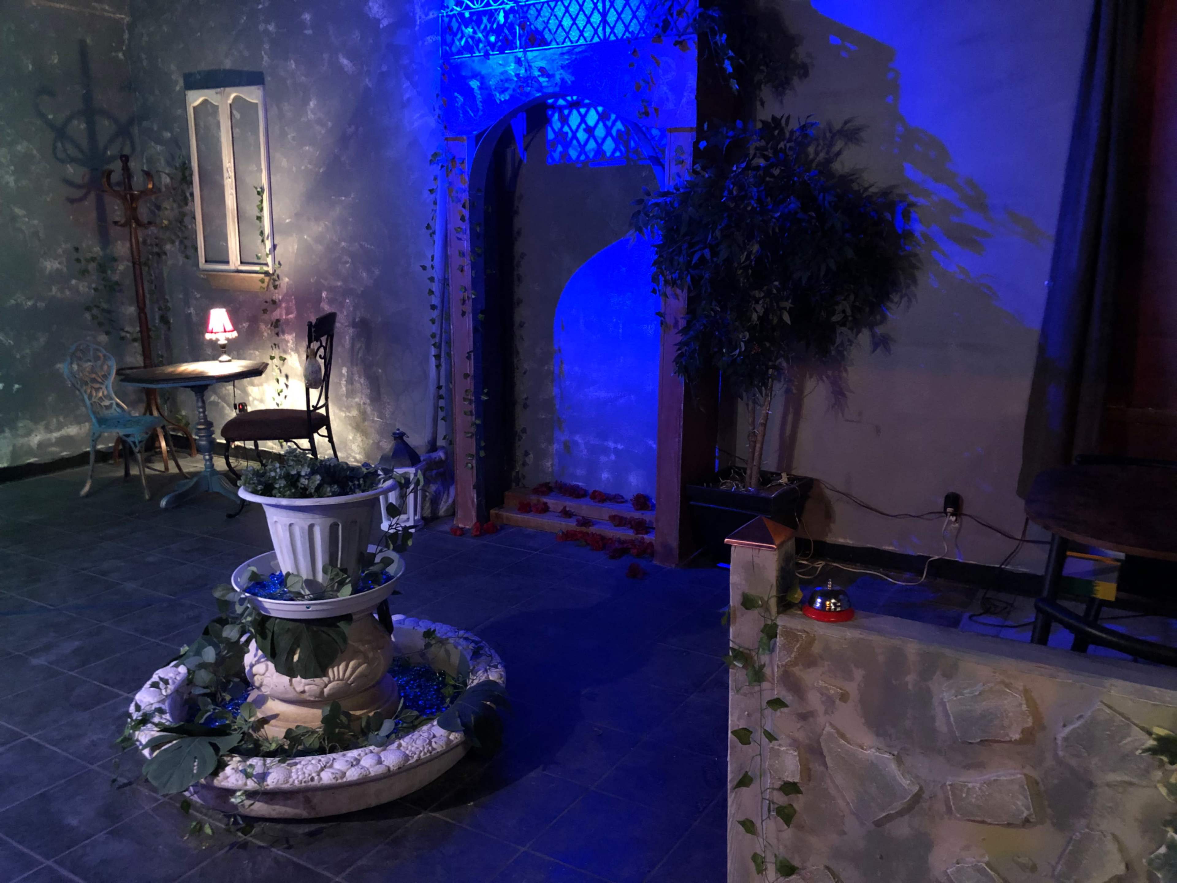 The scene features a dimly lit room with a decorative fountain surrounded by plants, a small table with a lamp, and an archway adorned with blue lighting.