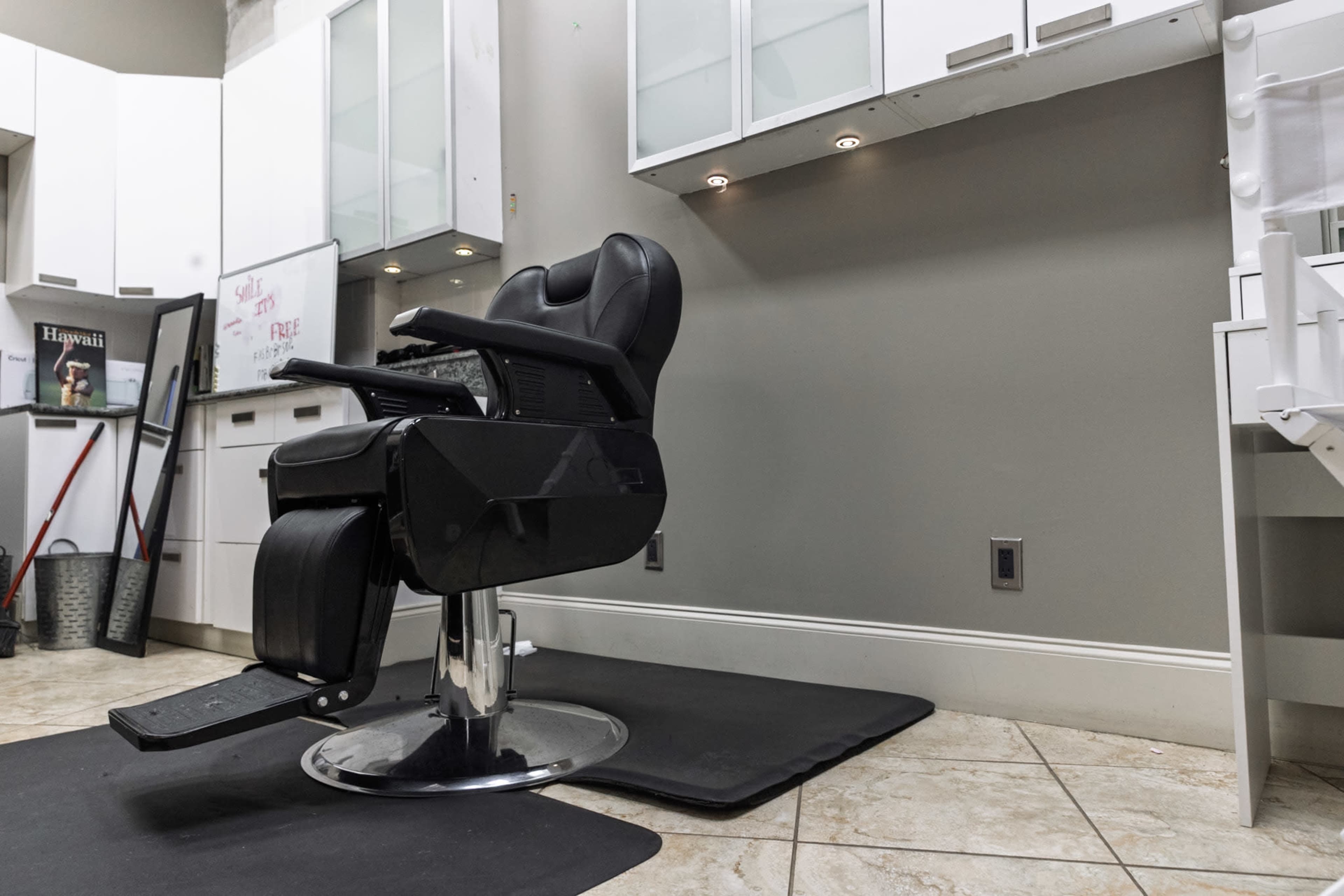 The image shows a black salon chair positioned on a mat in a modern hair salon with sleek cabinetry and a mirror in the background.