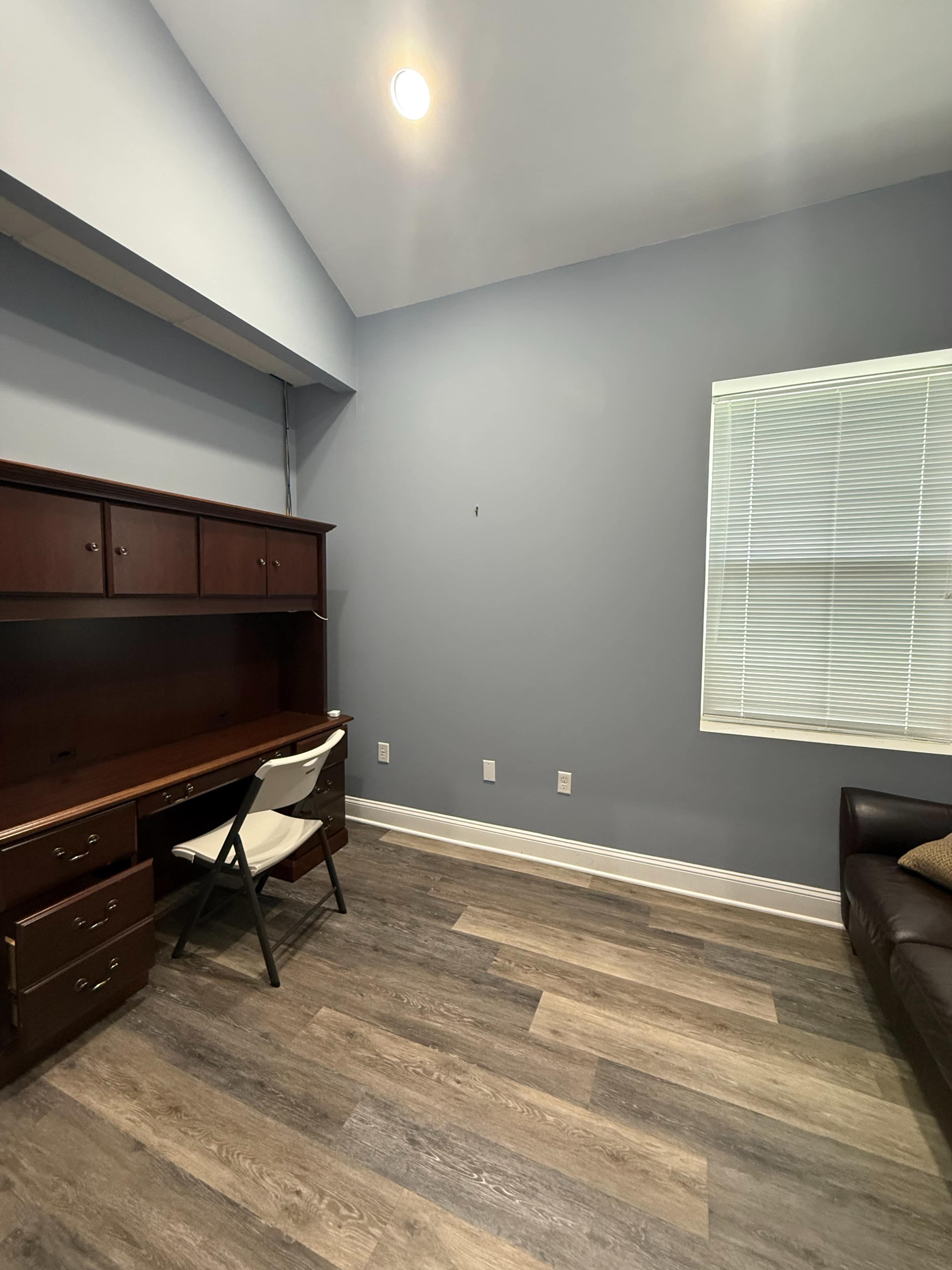 The image shows a room with a wooden desk and chair against a grey wall, featuring a window with blinds on the right side.