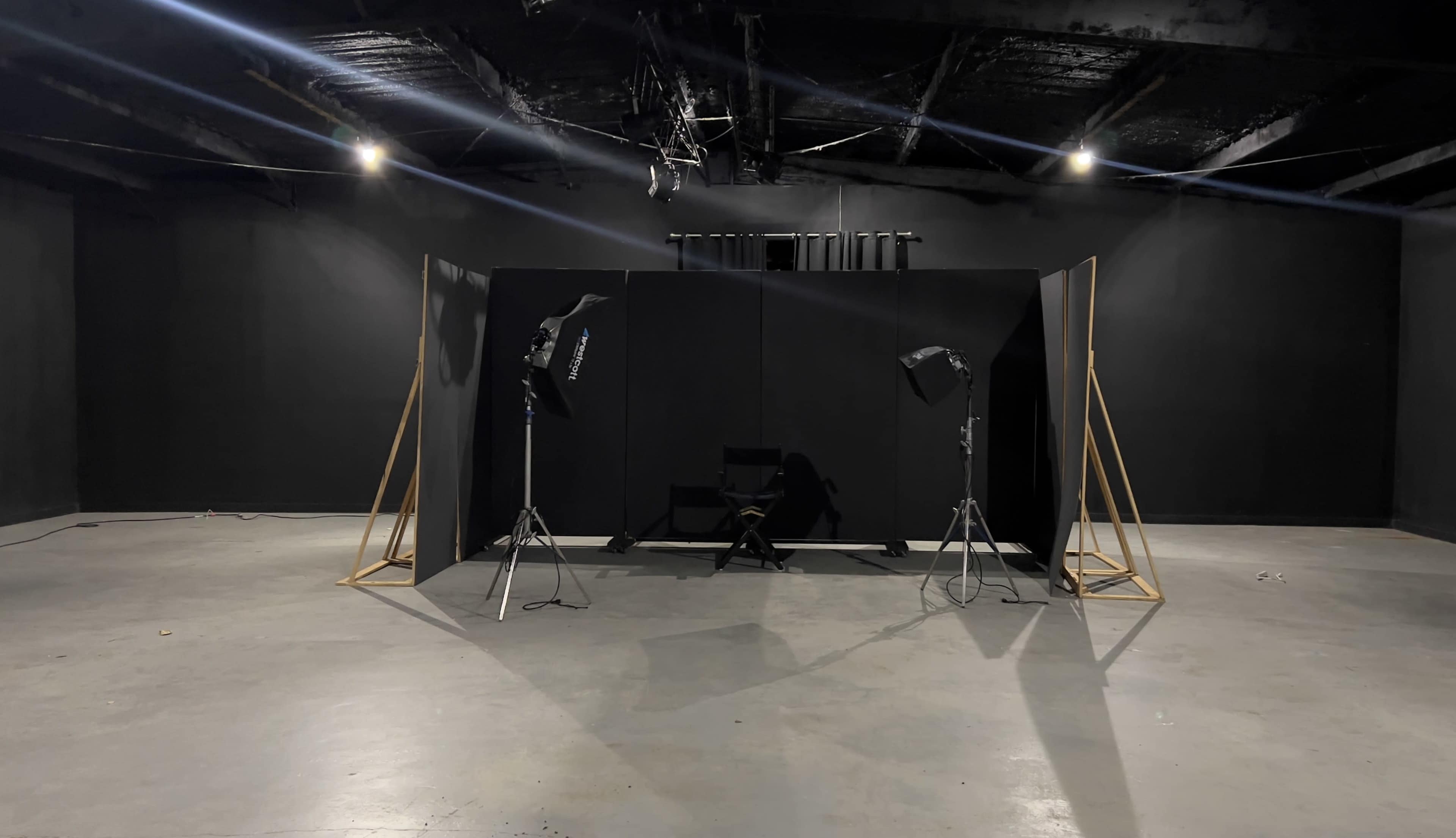 A minimalist photography studio setup with black walls, a director's chair, and lighting equipment positioned around a central backdrop.