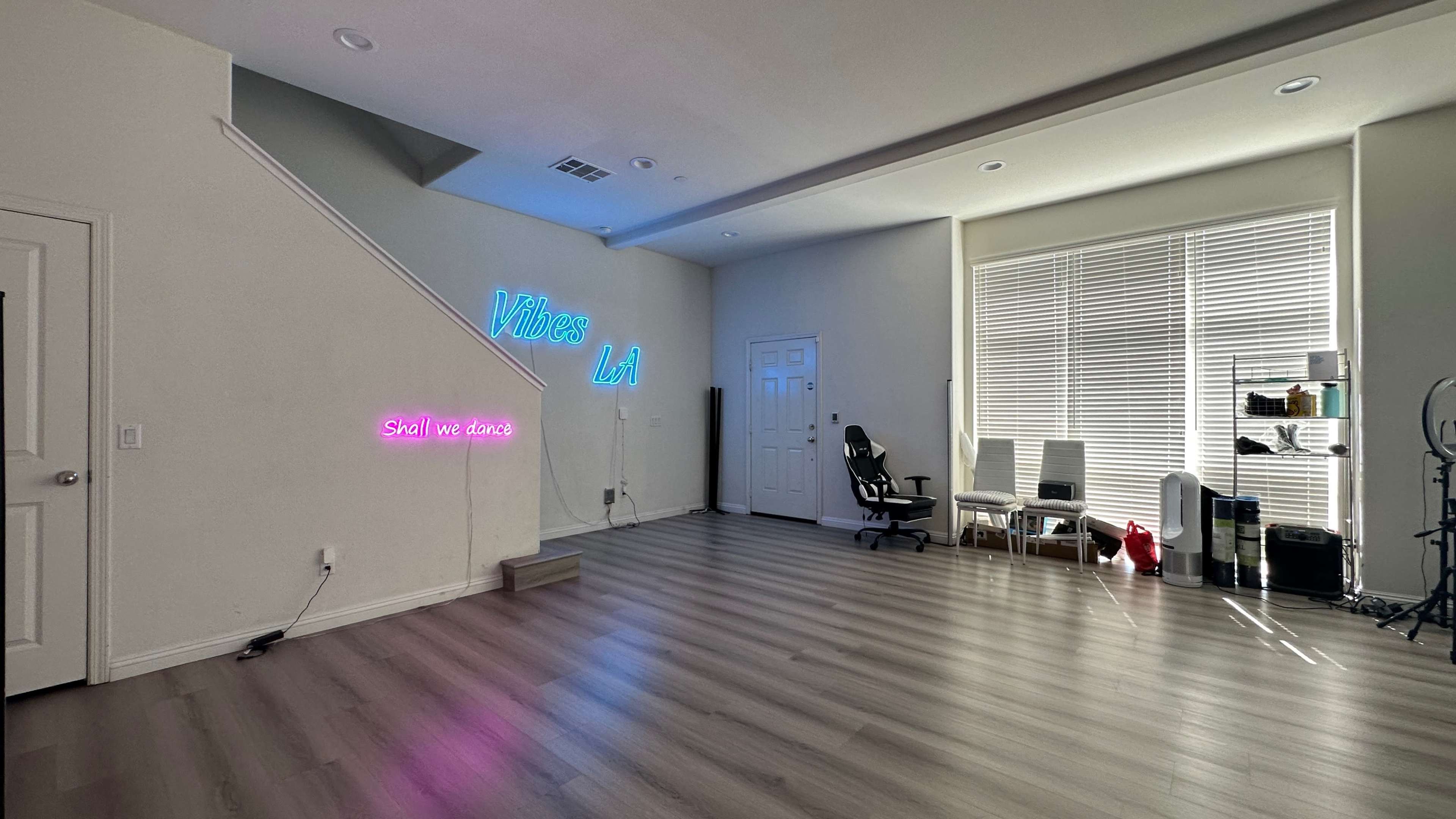 A spacious room features neon signs on the wall, a staircase, and a corner with a desk and chair near large windows.