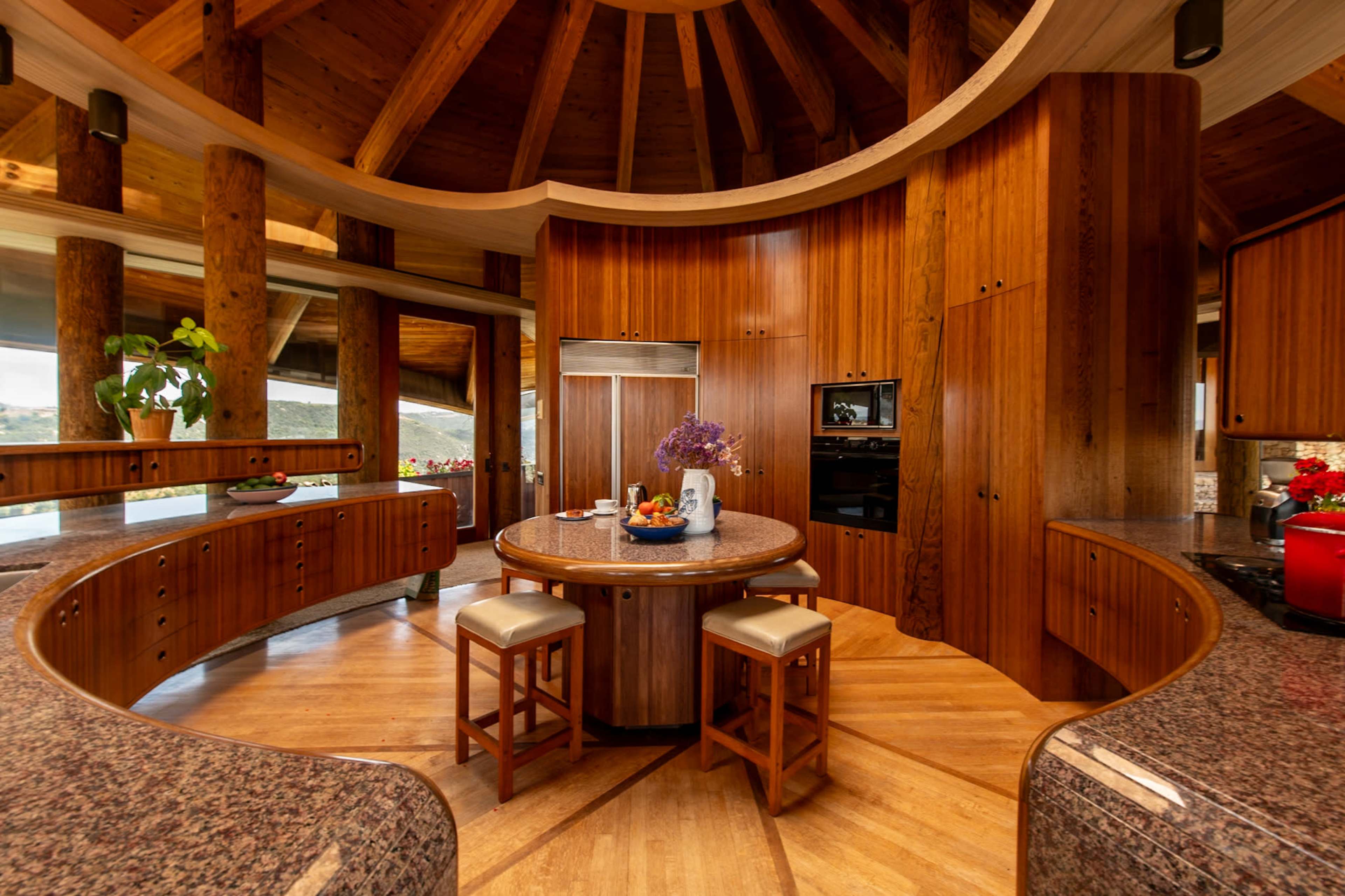 The image depicts a round kitchen featuring wooden cabinetry, a circular island with stools, and large windows allowing natural light.