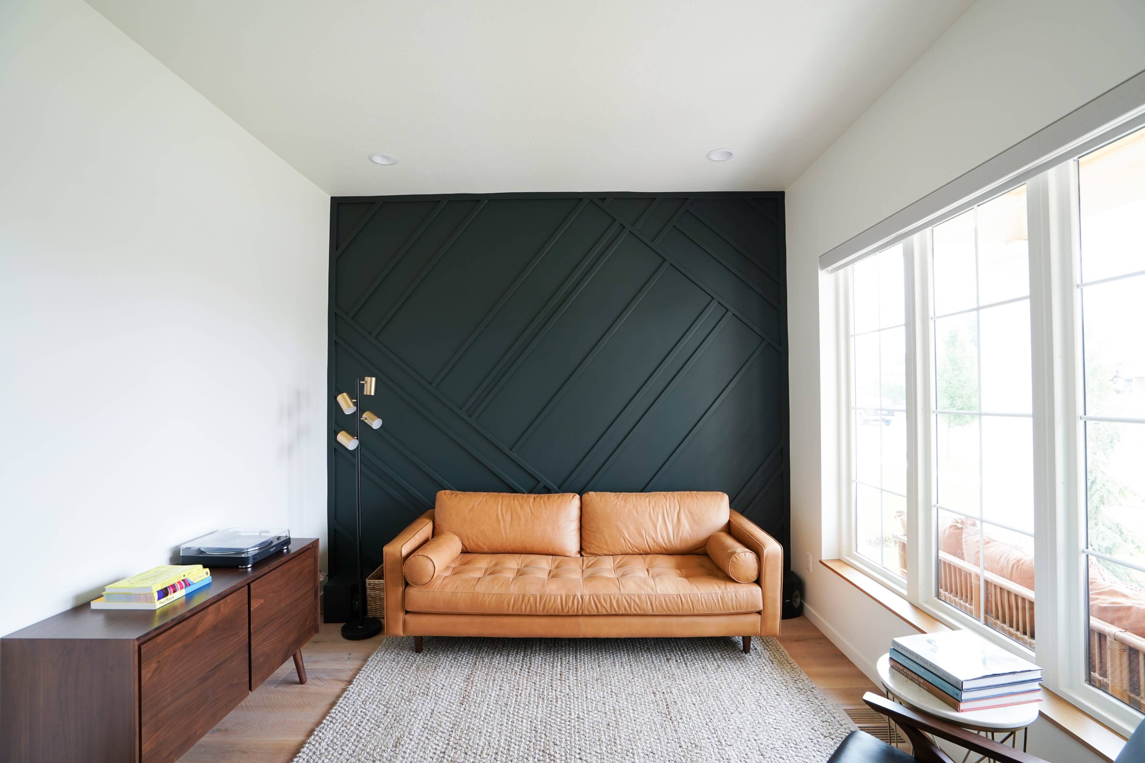 A leather couch is positioned against a geometric dark wall, with a large window and a wooden cabinet nearby.