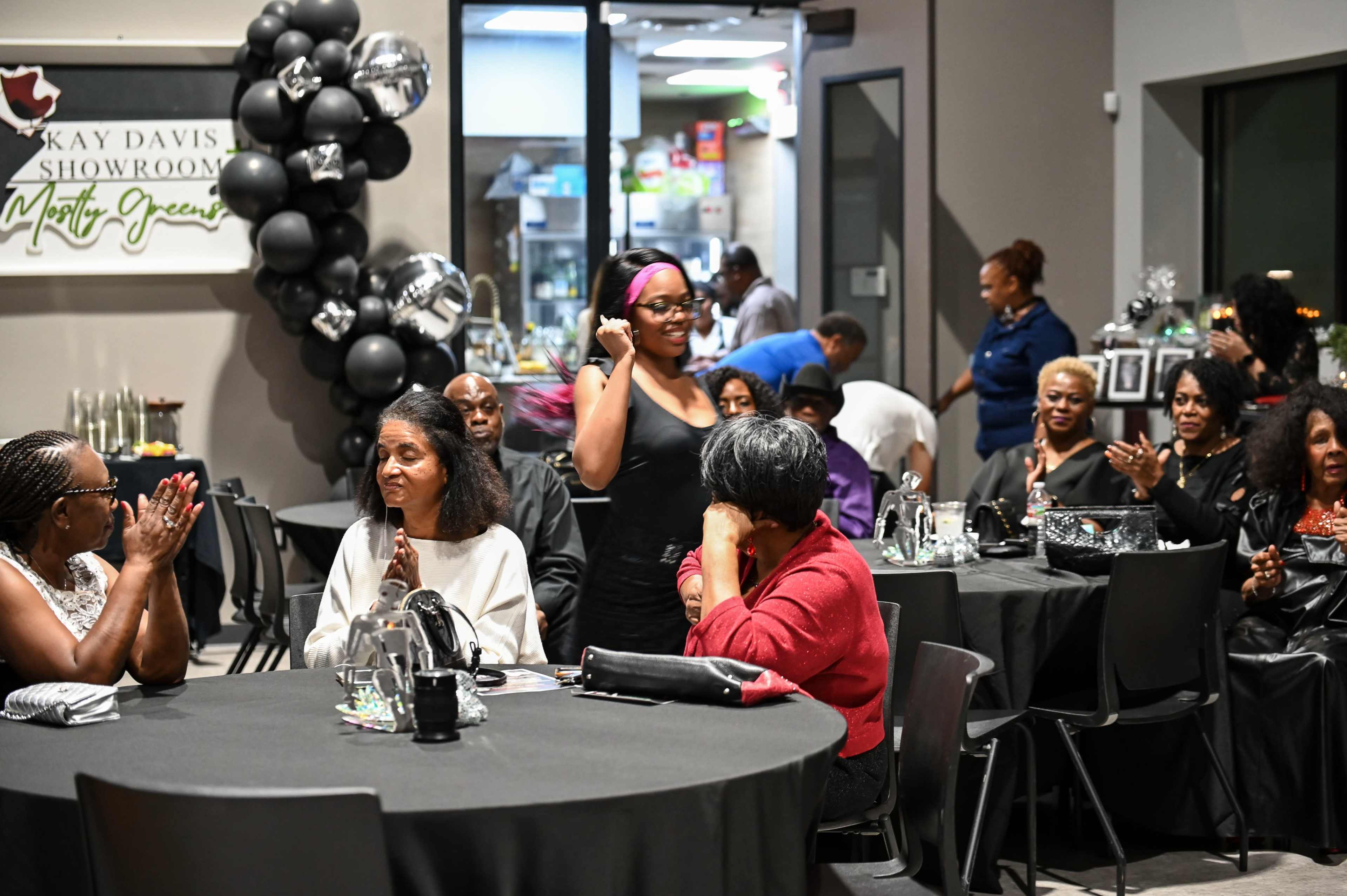 A group of people, mostly women, sits at tables in a showroom, while one woman walks through the space, and others are engaged in conversation or clapping.