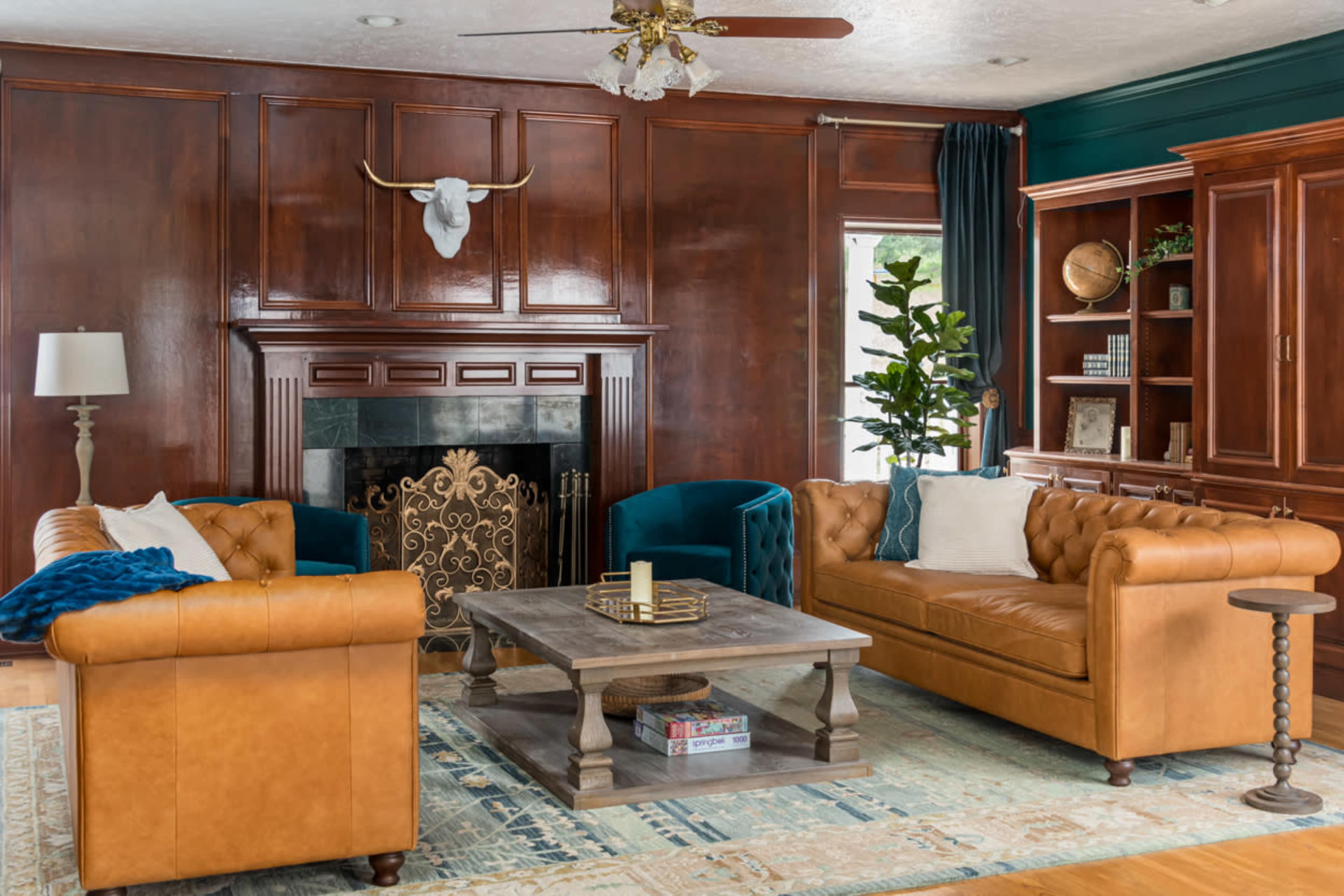 The image shows a styled living room featuring two leather sofas, a wooden coffee table, a decorative fireplace, and dark wood paneling on the walls.