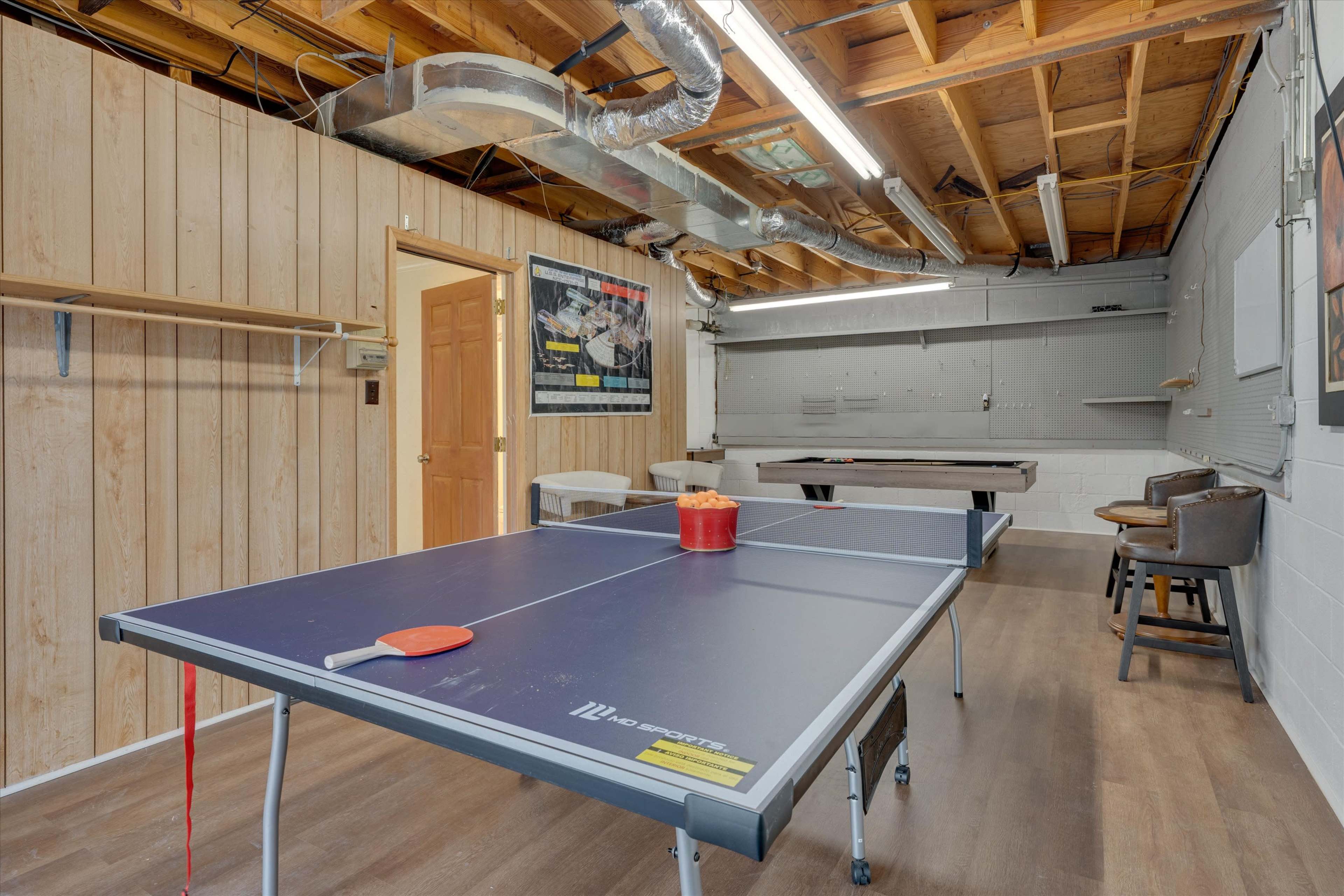 A basement recreation room featuring a ping pong table with a bucket of balls, wooden paneling on the walls, and a few stools against the back wall.