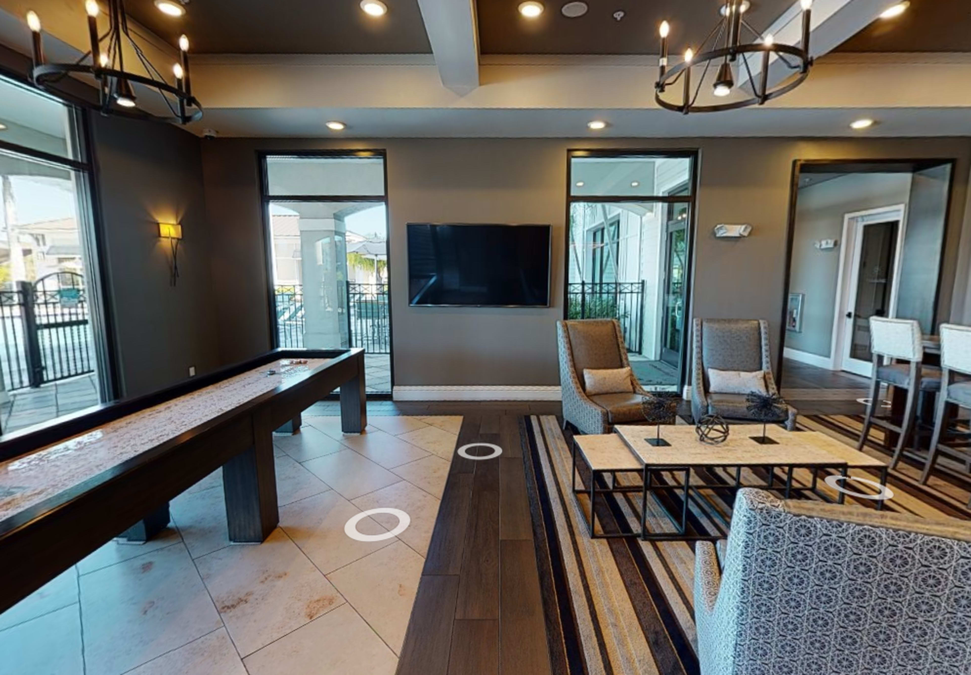 A modern lounge area with seating, a television, and a shuffleboard table, featuring large windows that allow natural light.