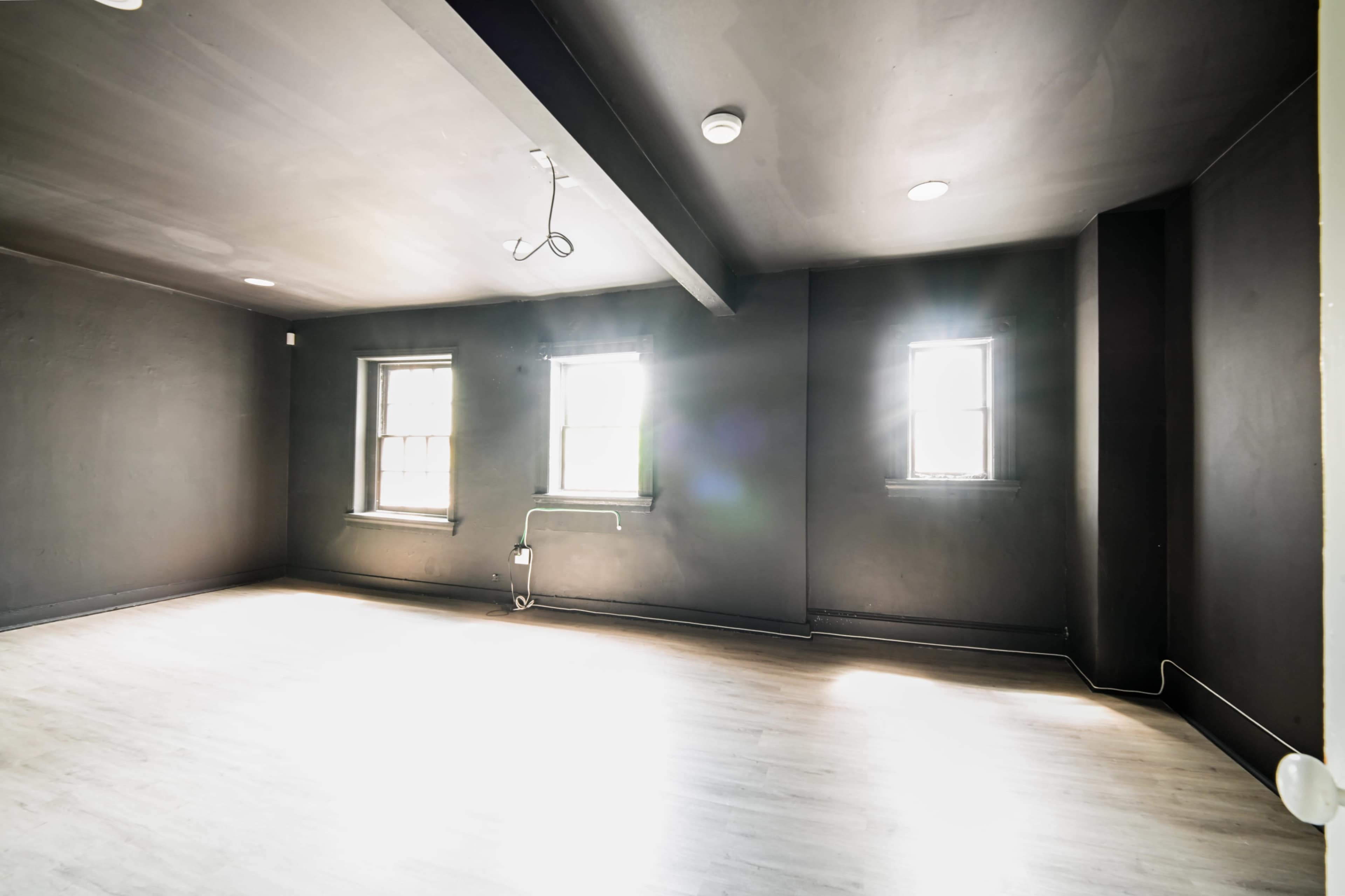 The image shows an empty room with black walls and two windows, featuring a minimalist design and wooden flooring.