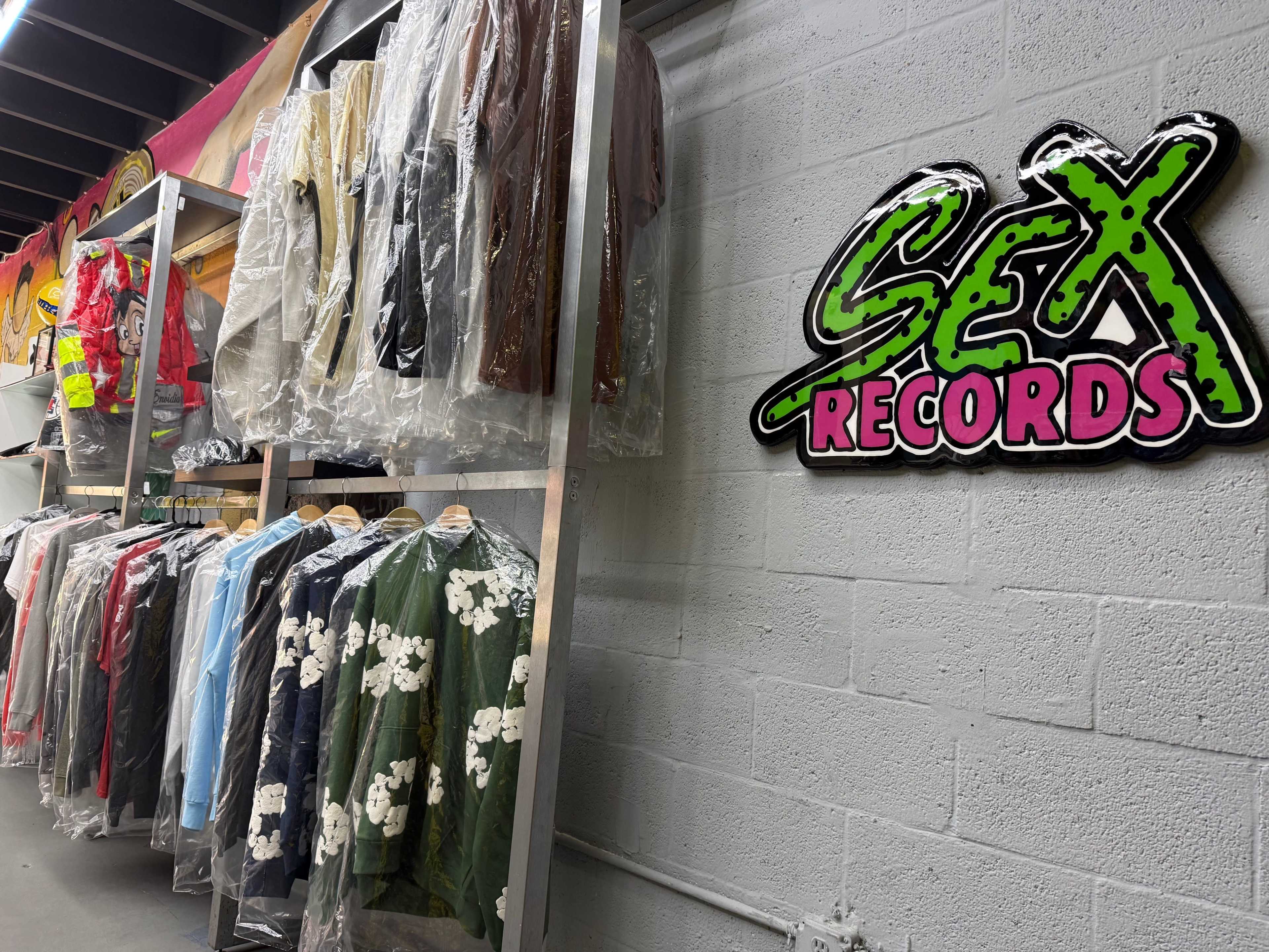 The image shows a clothing rack filled with various garments on hangers next to a brightly colored "SEX RECORDS" sign mounted on the wall.