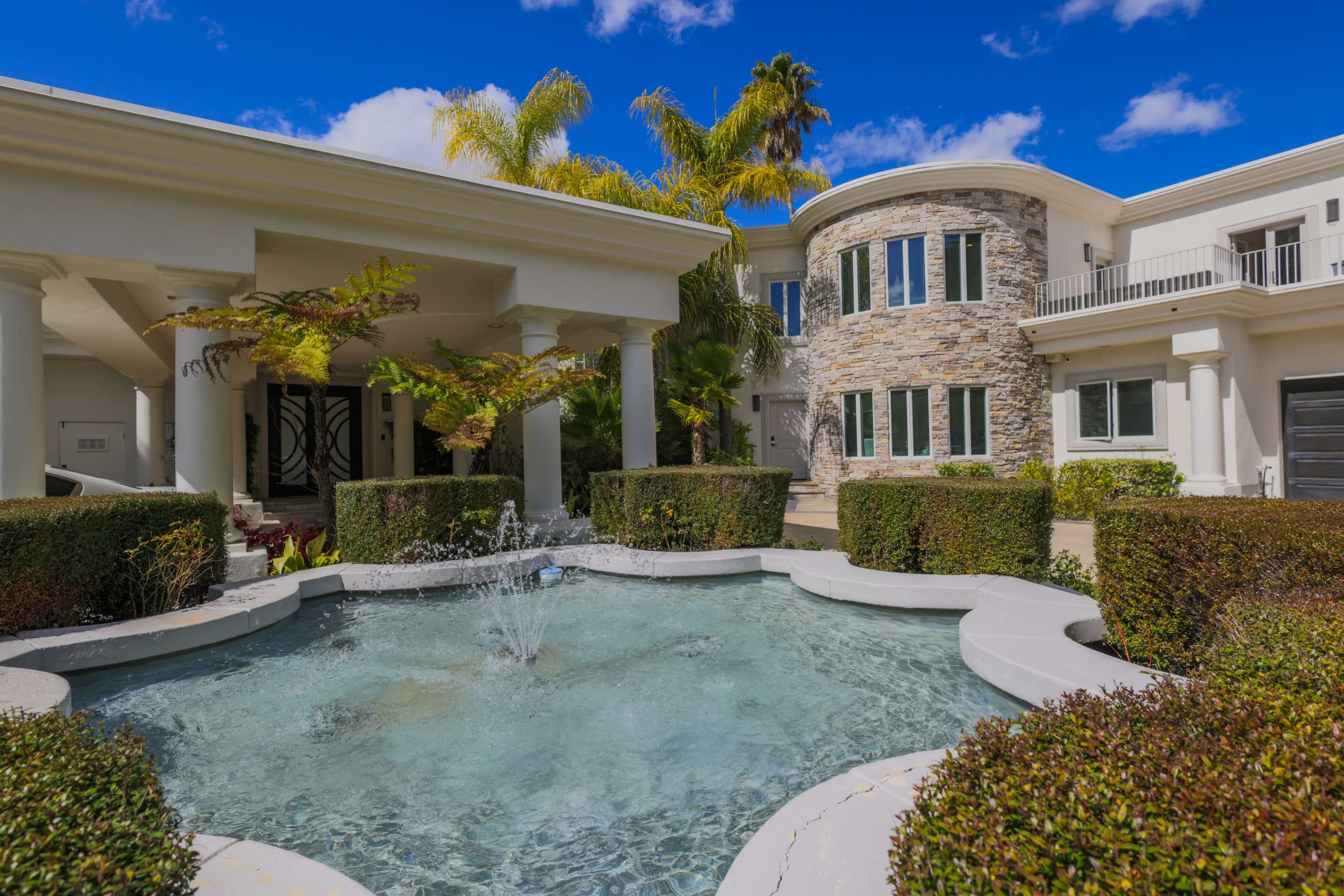 A large house with stone and stucco exterior, featuring a fountain surrounded by neatly trimmed hedges in the front courtyard.