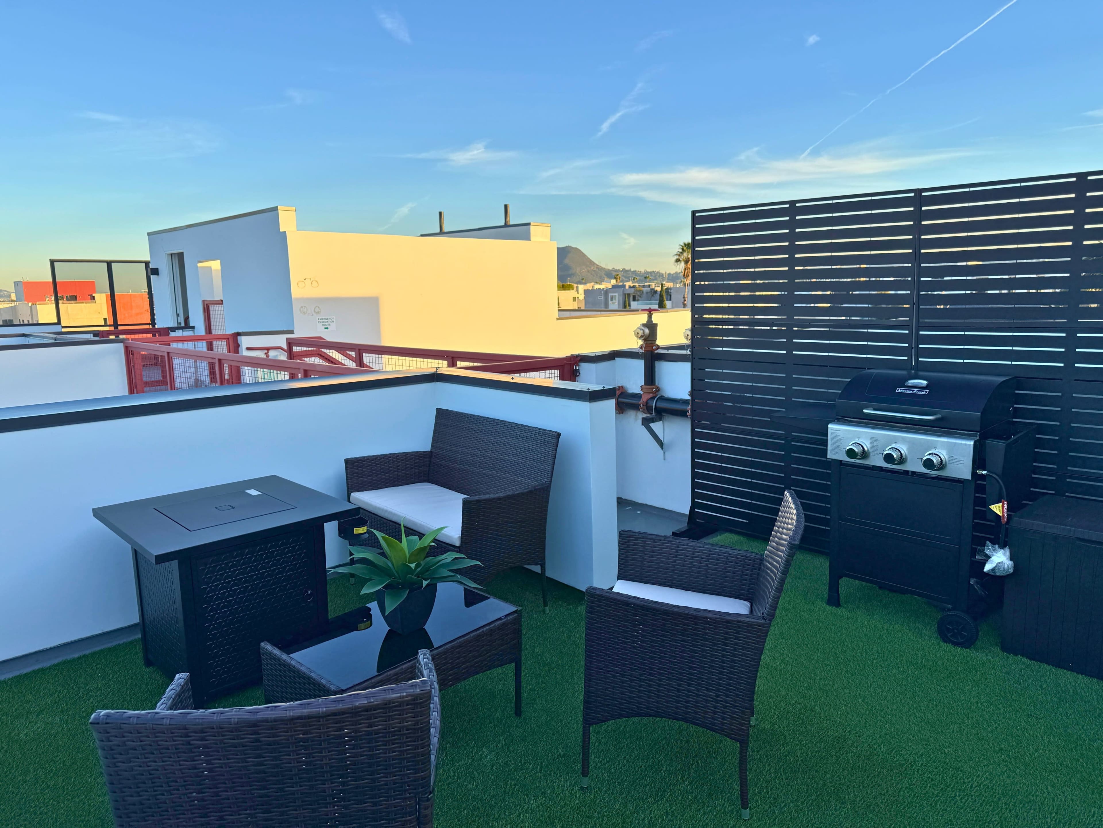 The image shows a rooftop terrace with wicker furniture, a small table, a BBQ grill, and a view of distant mountains against a clear blue sky.