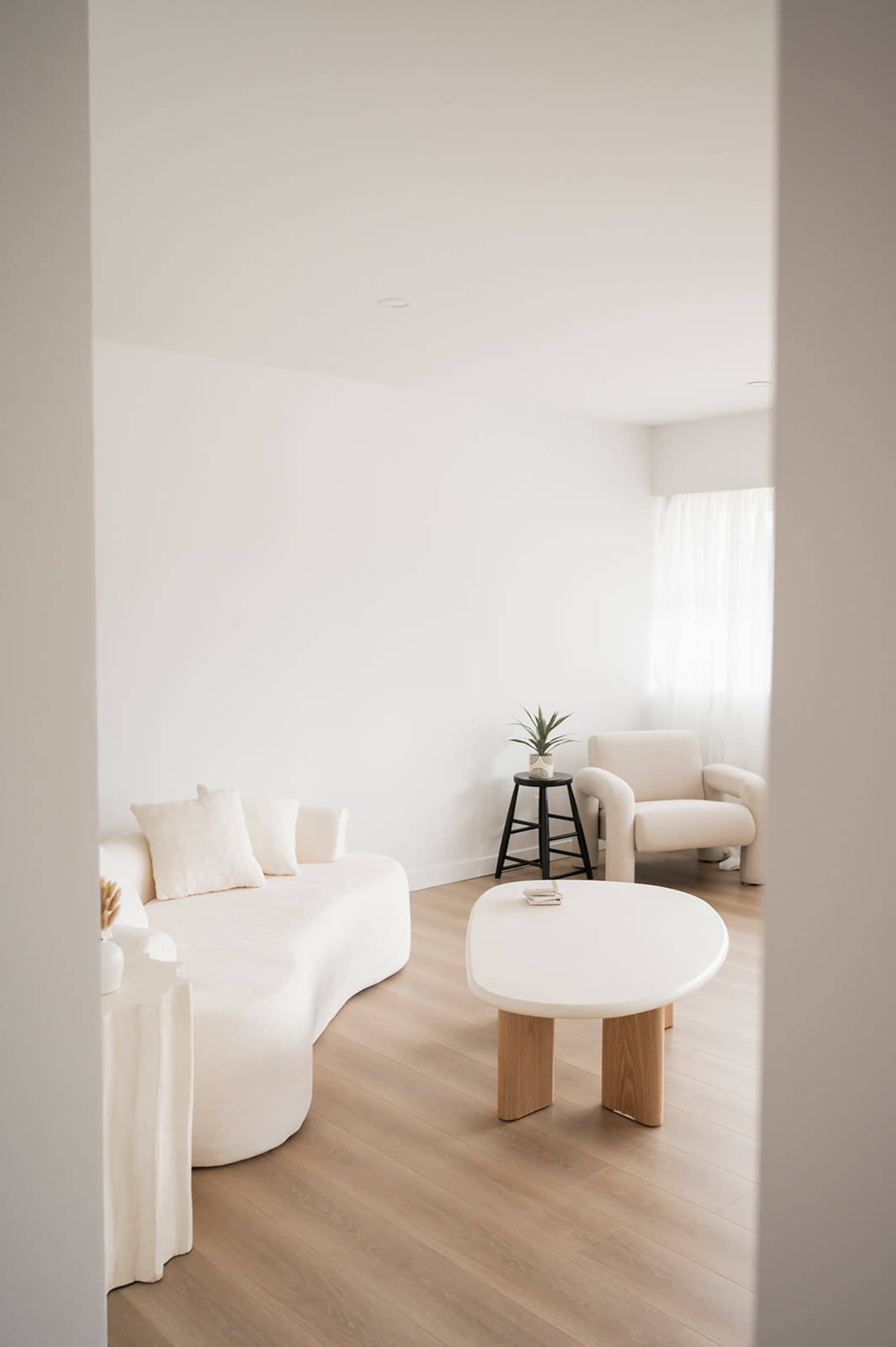 The image shows a modern, minimalist living room featuring a curved white sofa, a round coffee table with wooden legs, and a cozy armchair near a window with sheer white curtains.