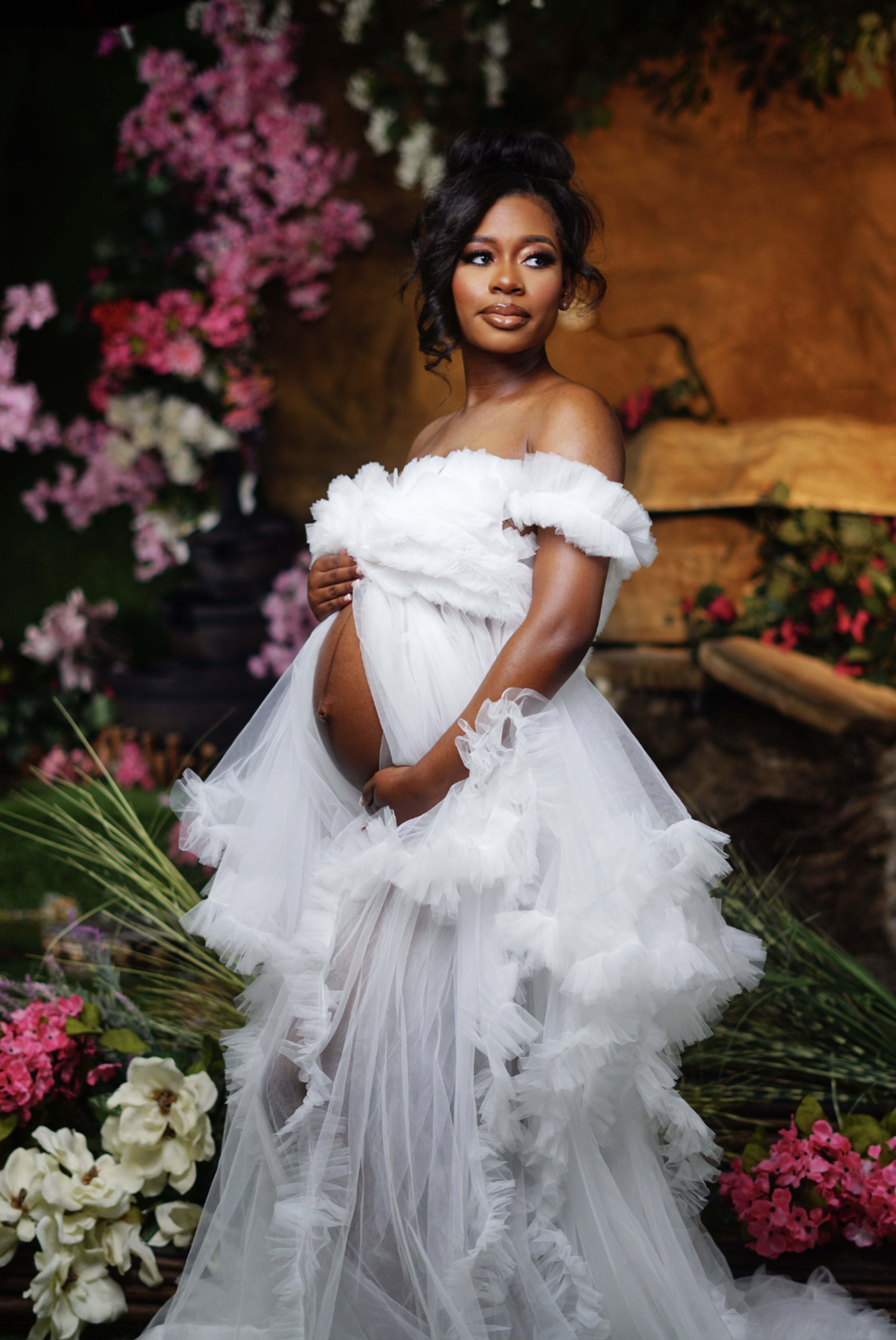 A pregnant woman in a white, ruffled gown stands gracefully among pink and white flowers.
