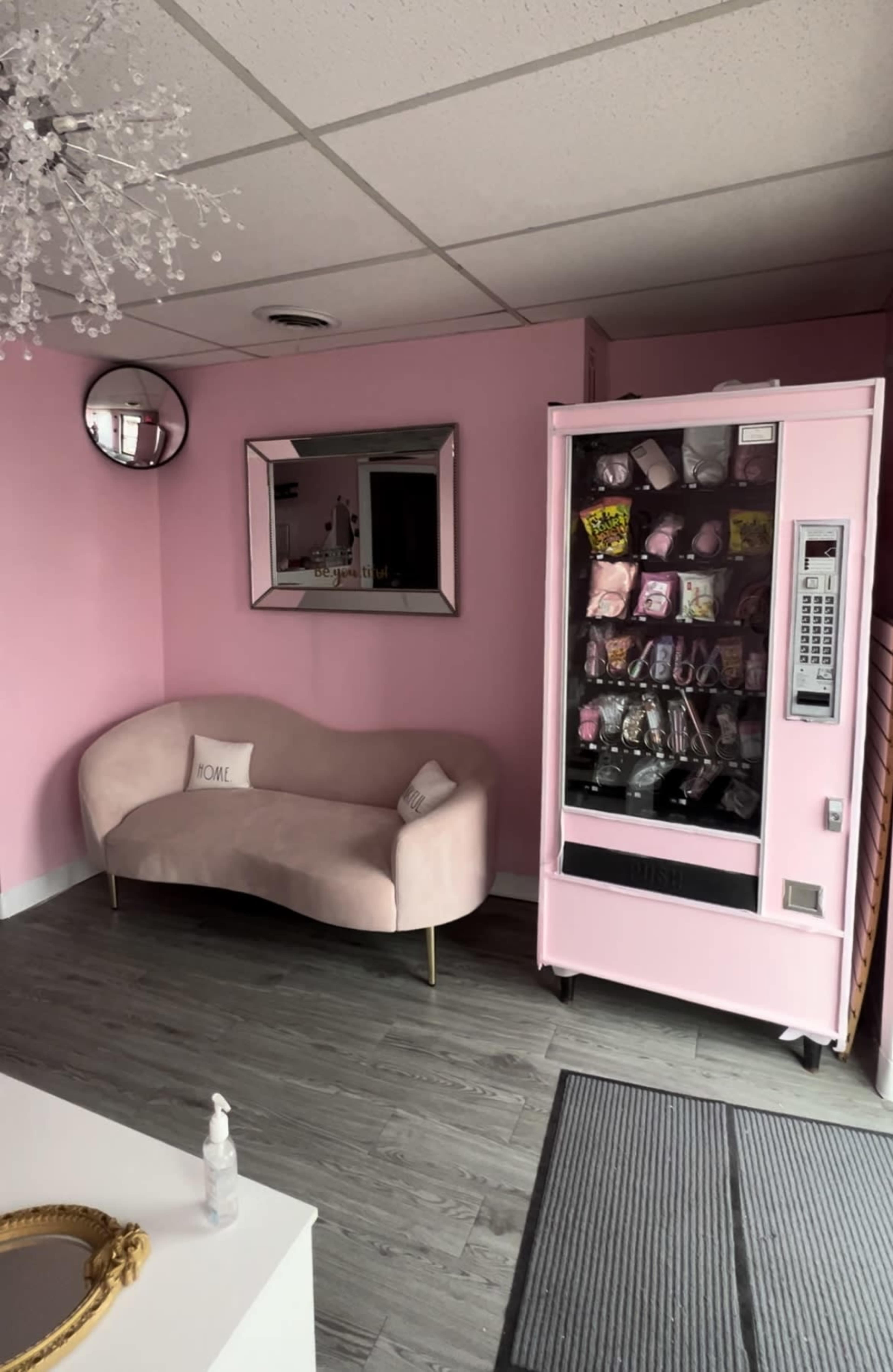 The image shows a small, pink-themed room with a curved seating area, a large mirror, and a vending machine filled with assorted snacks and products.