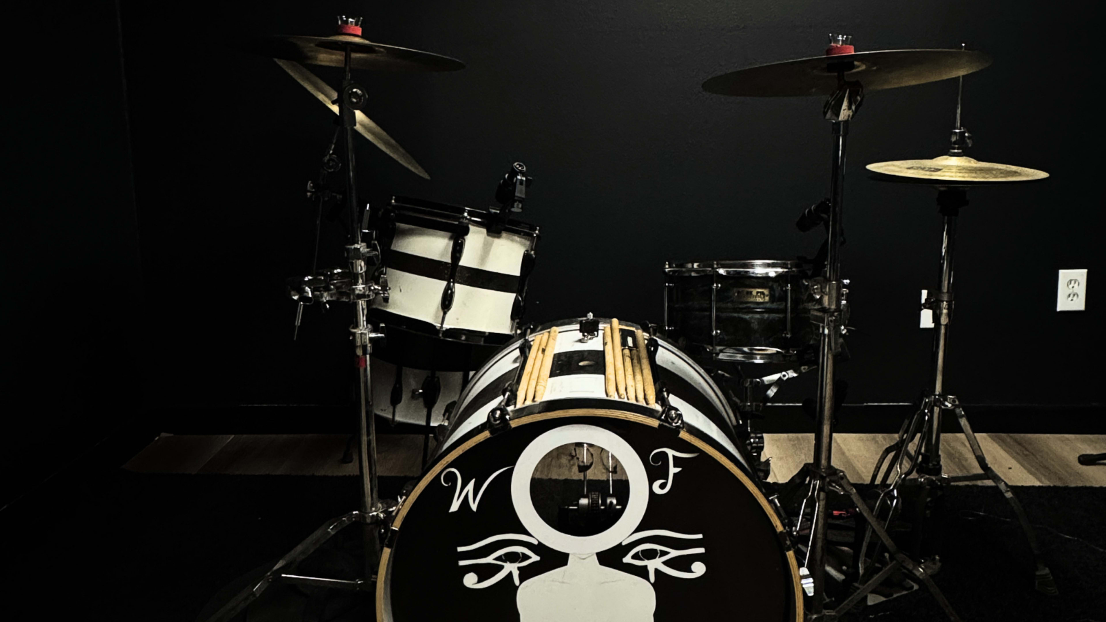 The image shows a drum set with a bass drum featuring a white logo, accompanied by a snare drum and cymbals, all set against a black background.