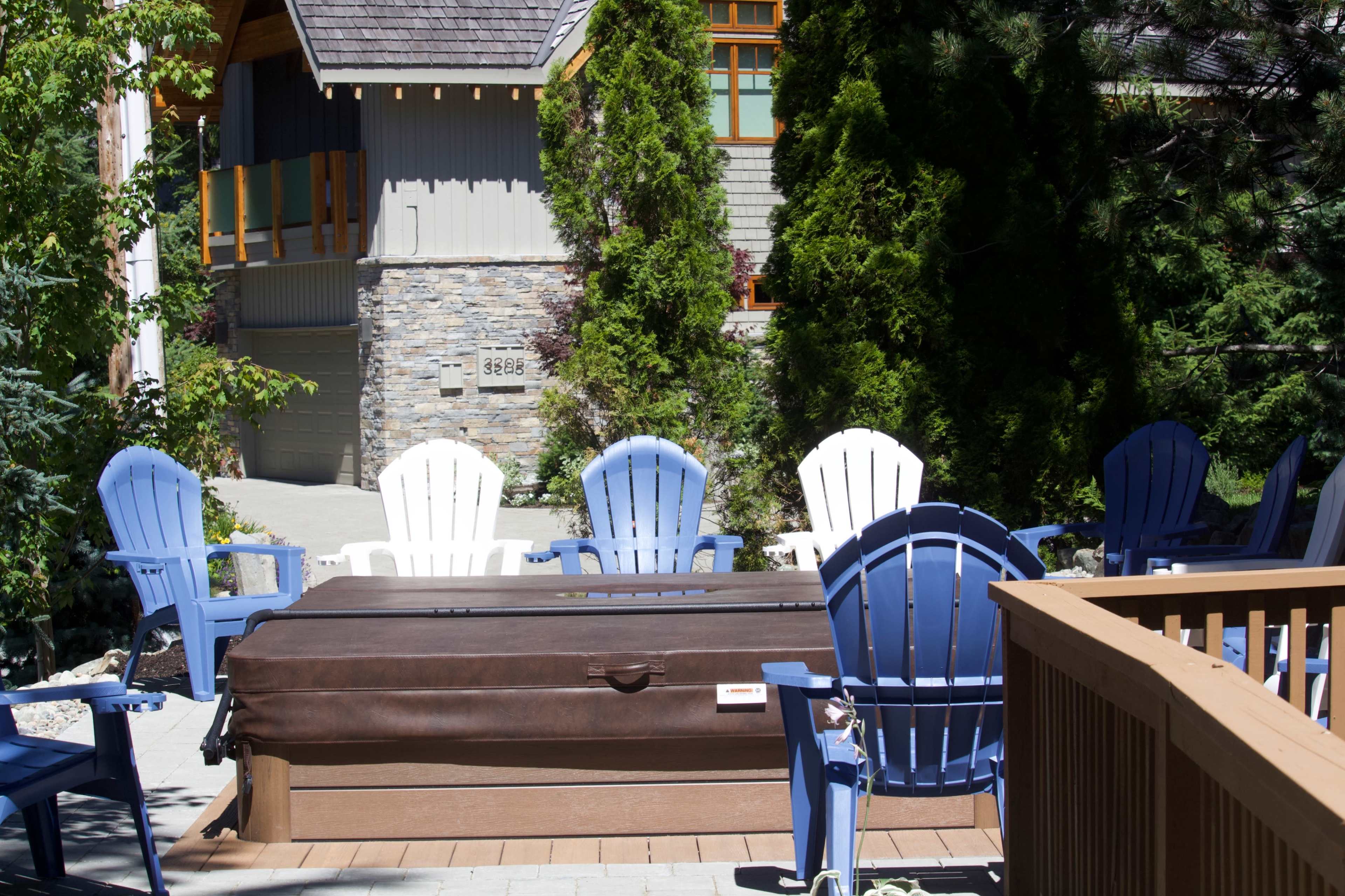 A row of blue and white Adirondack chairs surrounds a hot tub on a patio near a stone-faced house.