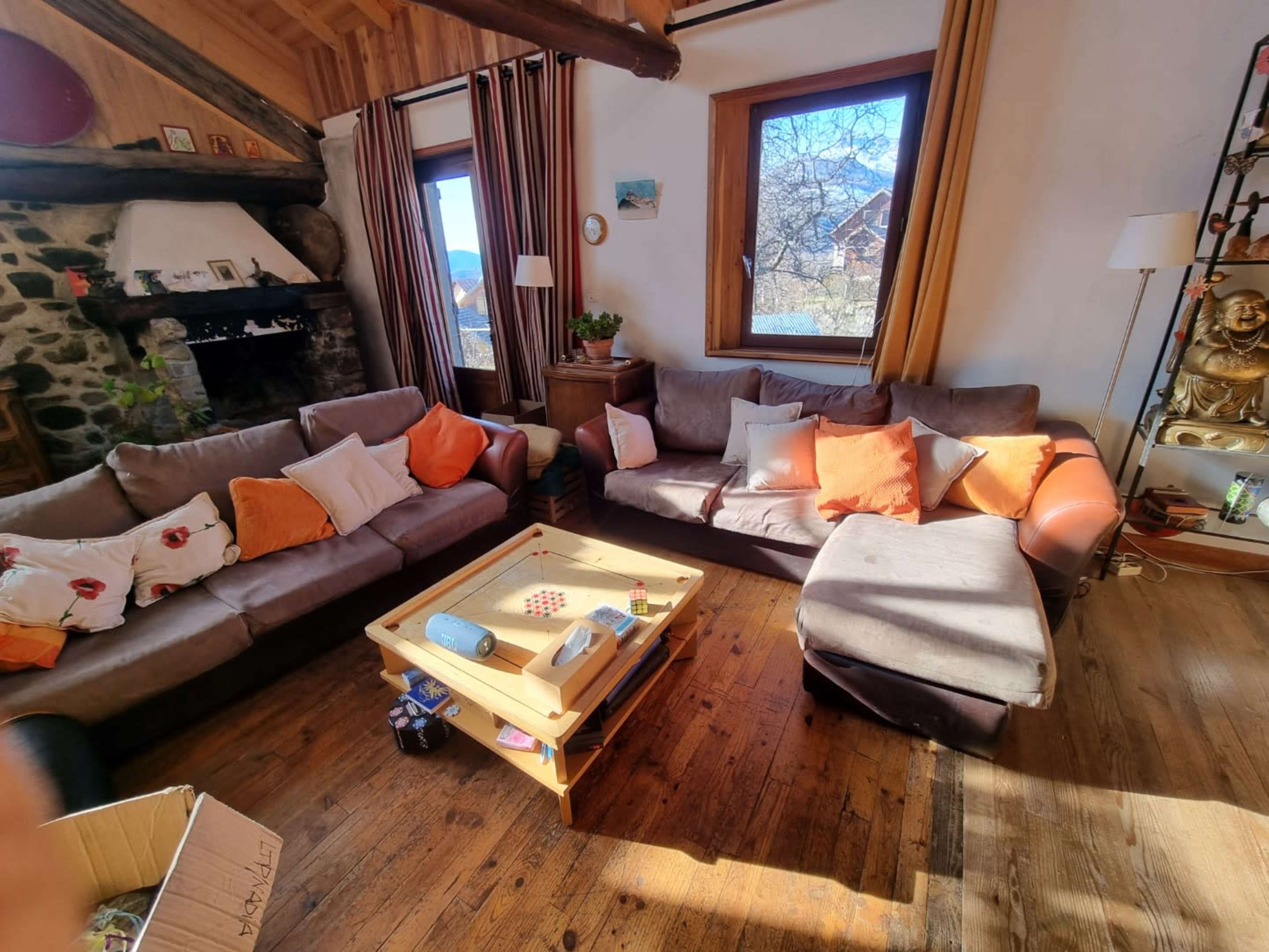 The scene shows a cozy living room with a large sectional sofa adorned with colorful cushions, a wooden coffee table in the center, and large windows allowing natural light to enter.